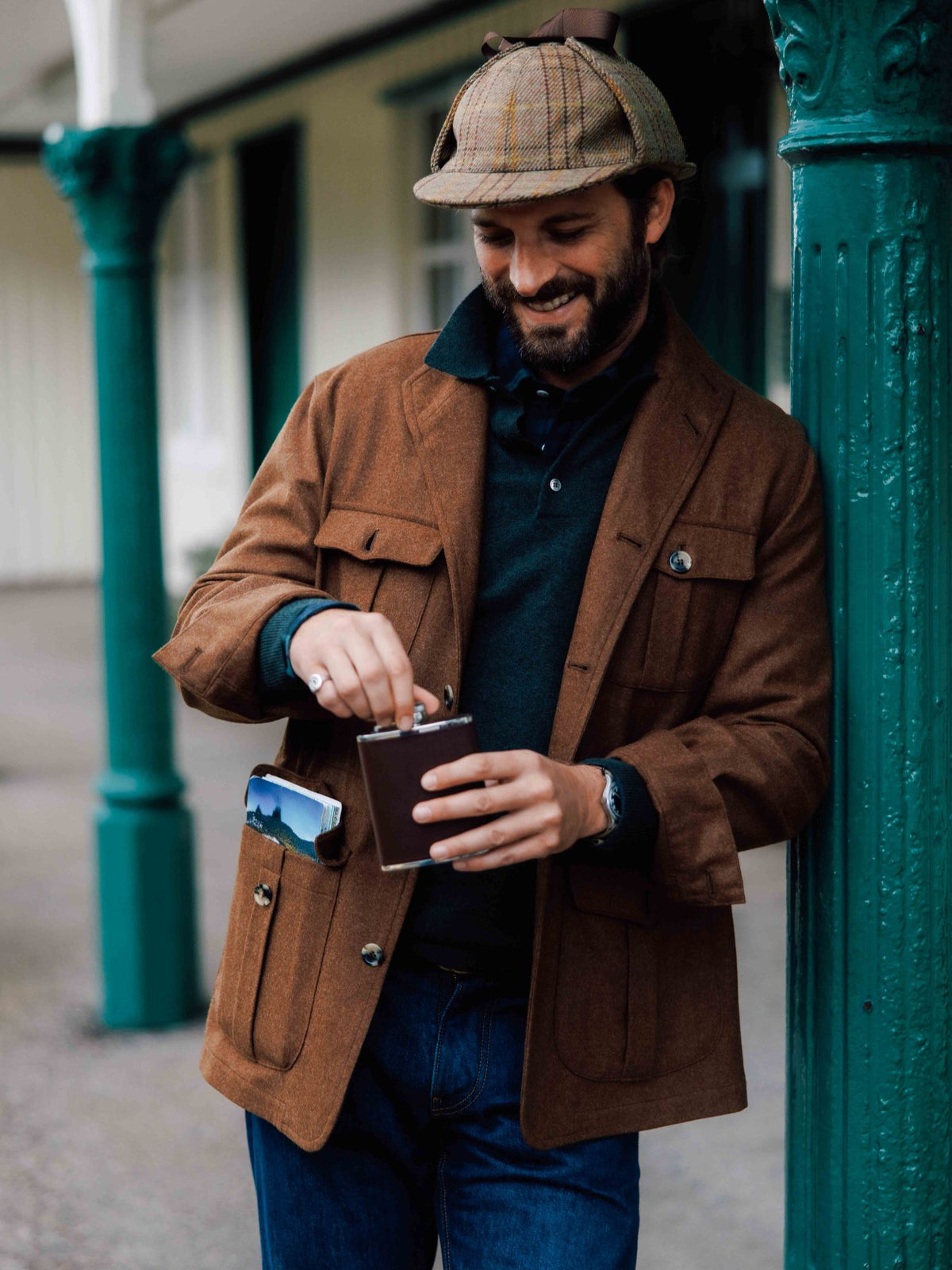 A man in a Campbell's of Beauly Field Teba Jacket, blue shirt, and flat cap smiles while holding a metal flask. He stands casually outdoors against a green pillar, with a phone visible in his jacket pocket.