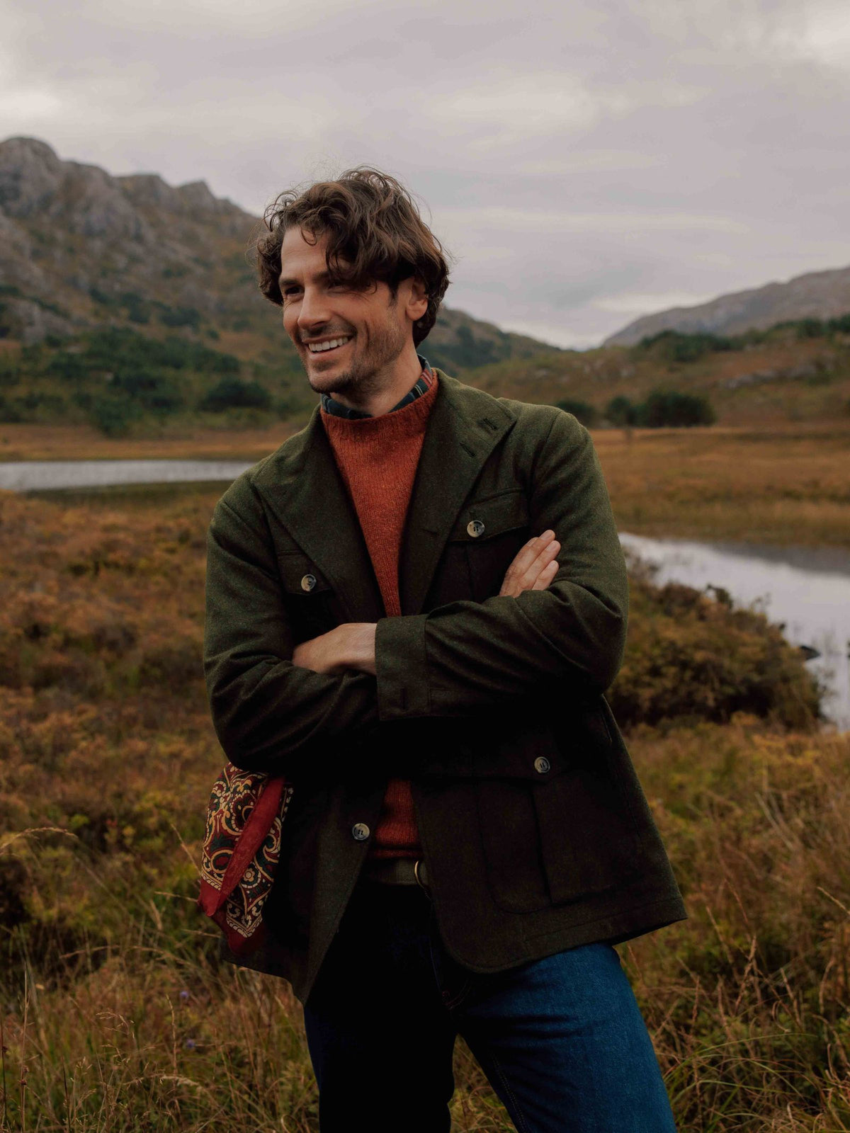 A man with wavy hair stands outdoors in a grassy setting, smiling with arms crossed. He wears the Campbells of Beauly Field Teba Jacket, a rust-red sweater, jeans, and carries a patterned bag over his shoulder.