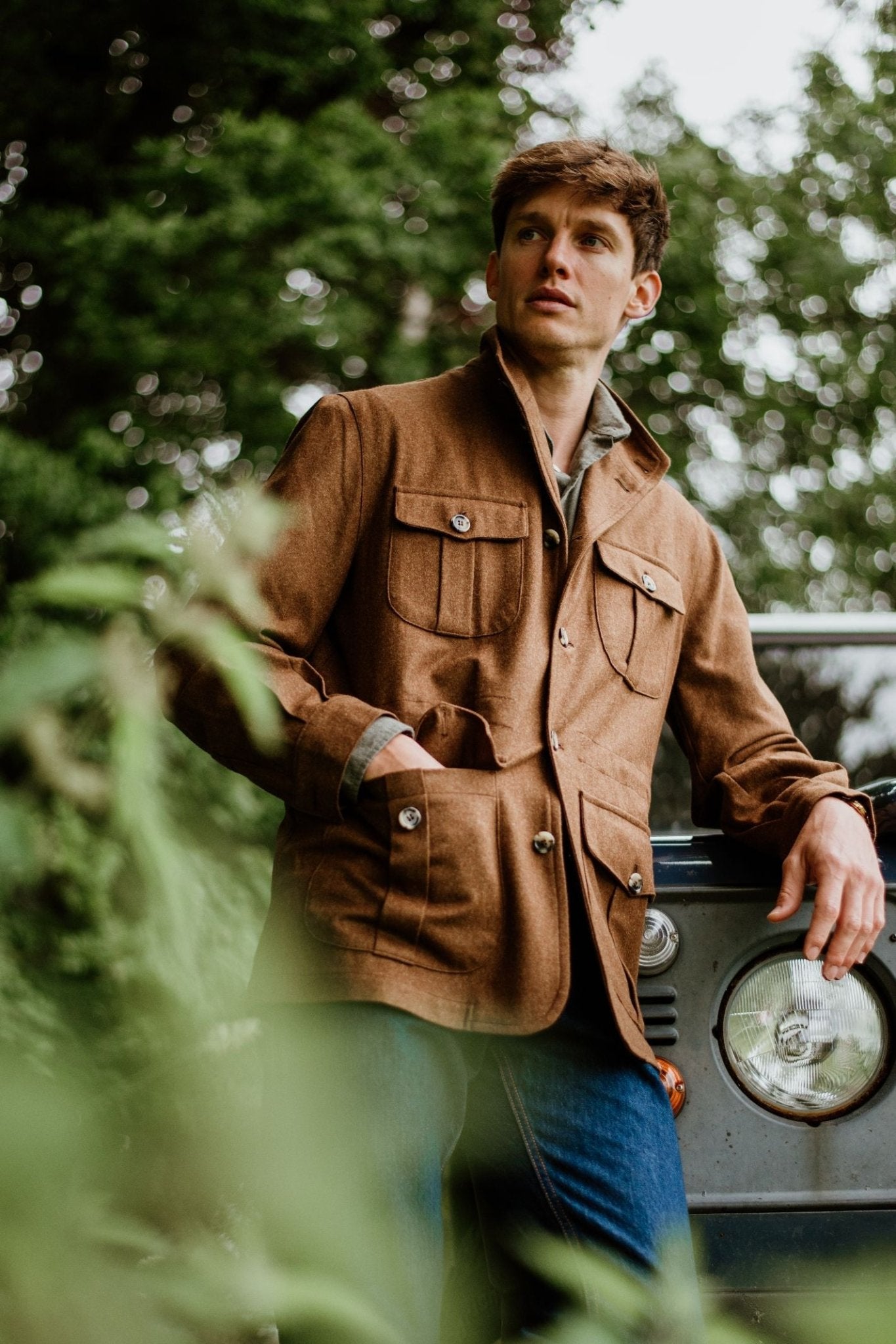 A man in a Campbell's of Beauly Field Teba Jacket and blue jeans leans against a vintage vehicle outdoors, surrounded by greenery, gazing thoughtfully into the distance.