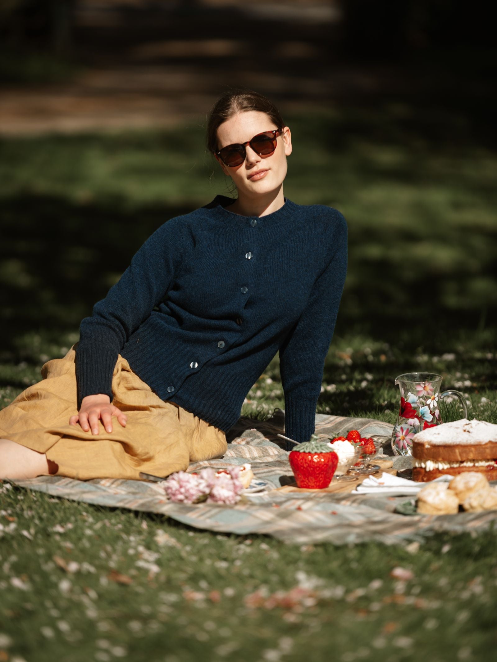 A woman in sunglasses and a Campbell's of Beauly Geelong Lambswool Crew Cardigan sits on a picnic blanket outside, surrounded by grass and sunlight. On the blanket are a tan skirt, red apple, pastries, cake, and a pitcher of water.