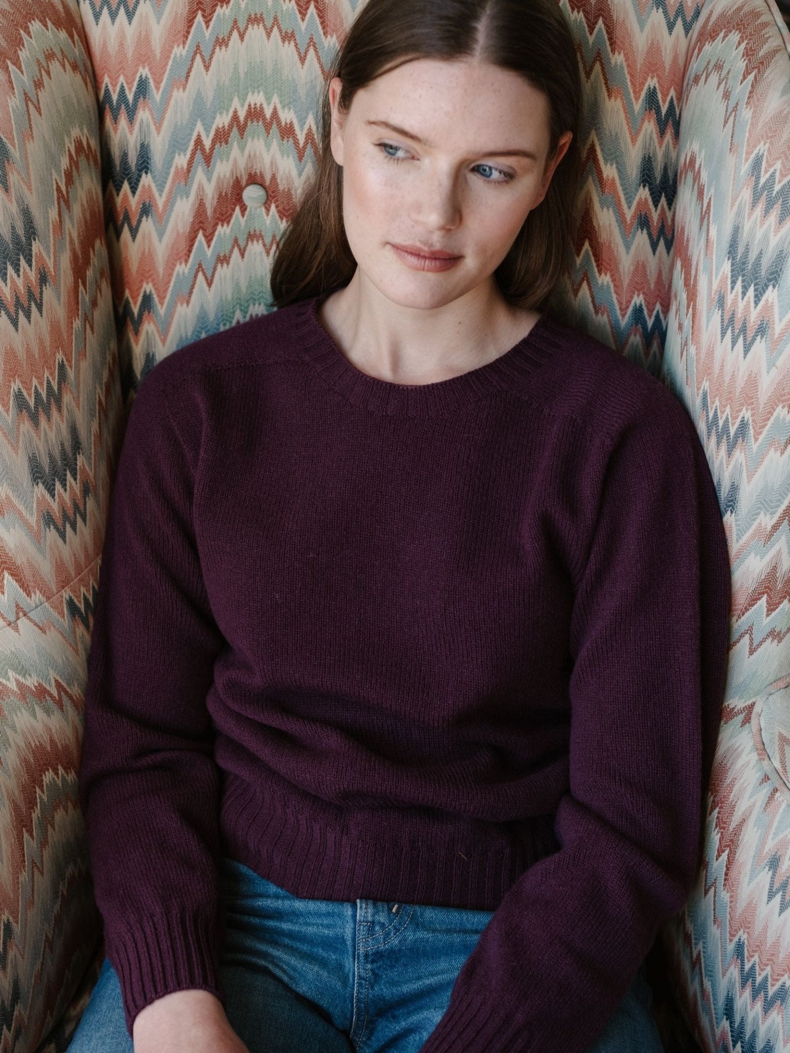 A woman with straight brown hair, wearing a maroon Campbell's of Beauly Geelong Lambswool Crew Neck Jumper and blue jeans, sits in a patterned armchair and gazes calmly to the side.