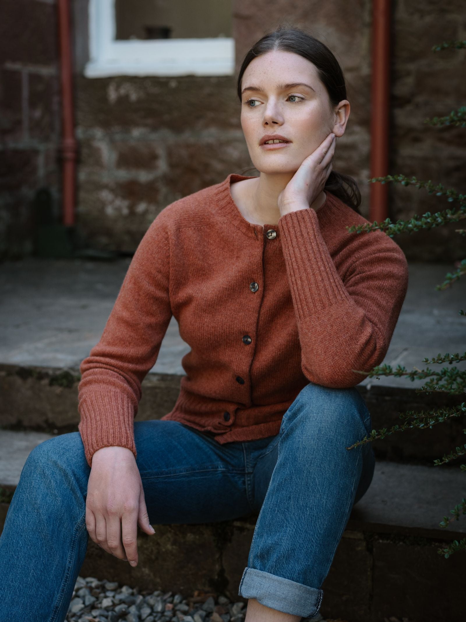 A woman with brown hair in a low ponytail, wearing Campbell's of Beauly’s Geelong Lambswool Crew Cardigan in Copper and blue jeans, sits on stone steps outside, resting her hand on her neck and gazing to the side thoughtfully.