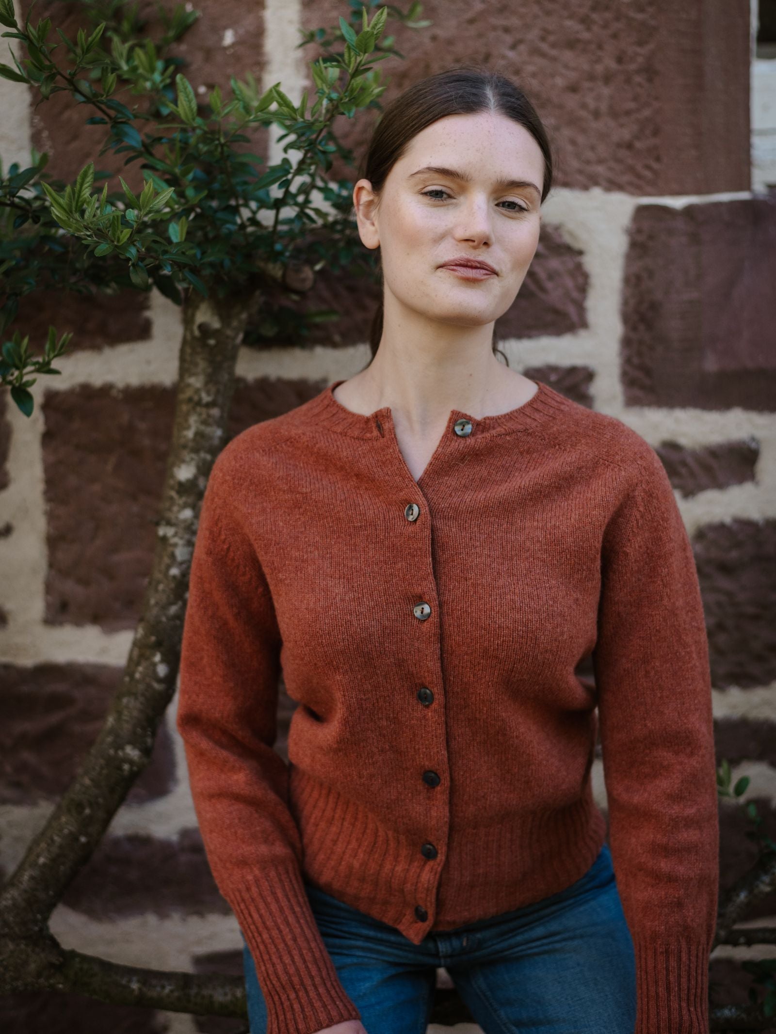 A woman with straight brown hair stands calmly before a stone wall, wearing Campbell's of Beauly's Geelong Lambswool Crew Cardigan in rust and blue jeans, with a leafy branch beside her.