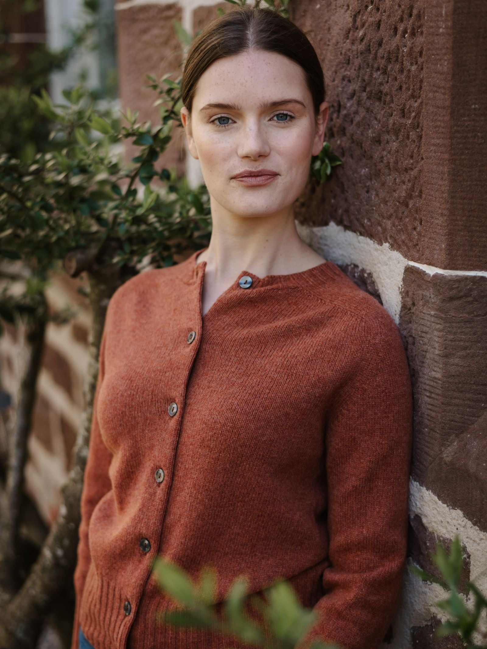 A woman with fair skin and brown hair tied back stands against a red brick wall, wearing a rust-colored Geelong Lambswool Crew Cardigan by Campbell's of Beauly. She looks at the camera neutrally, with greenery in the background.