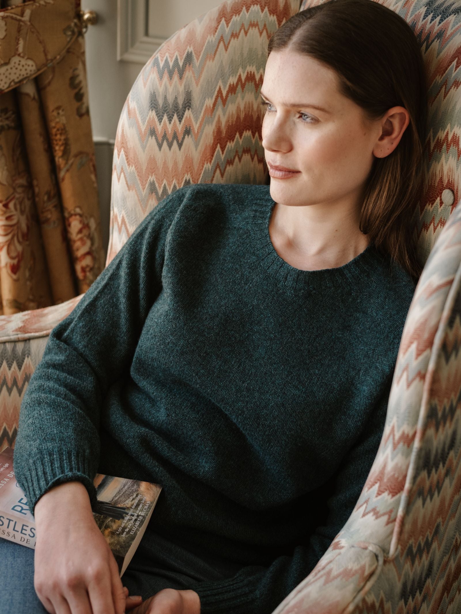 A woman with straight brown hair sits in a patterned armchair wearing a teal Campbell's of Beauly Geelong Lambswool Crew Neck Jumper, holding a book and gazing thoughtfully out of the frame.
