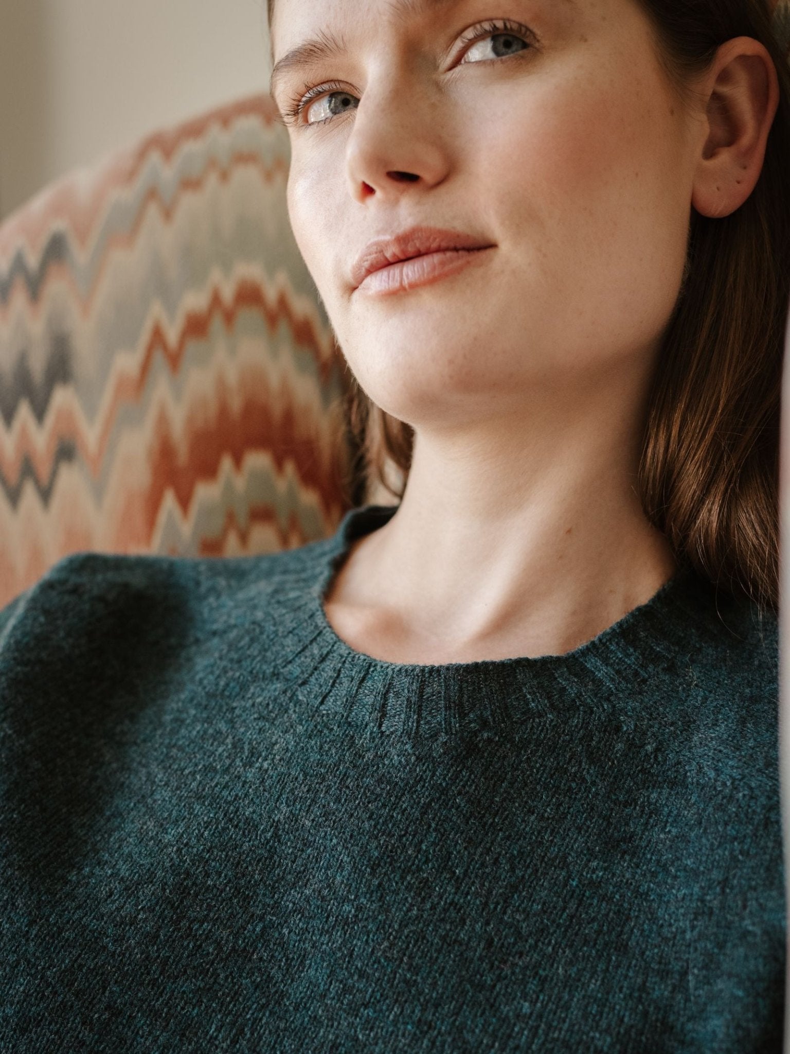 A woman with fair skin and brown hair sits on a patterned chair, wearing the Campbell's of Beauly Geelong Lambswool Crew Neck Jumper in dark green, and gazes slightly upward with a soft, relaxed expression.