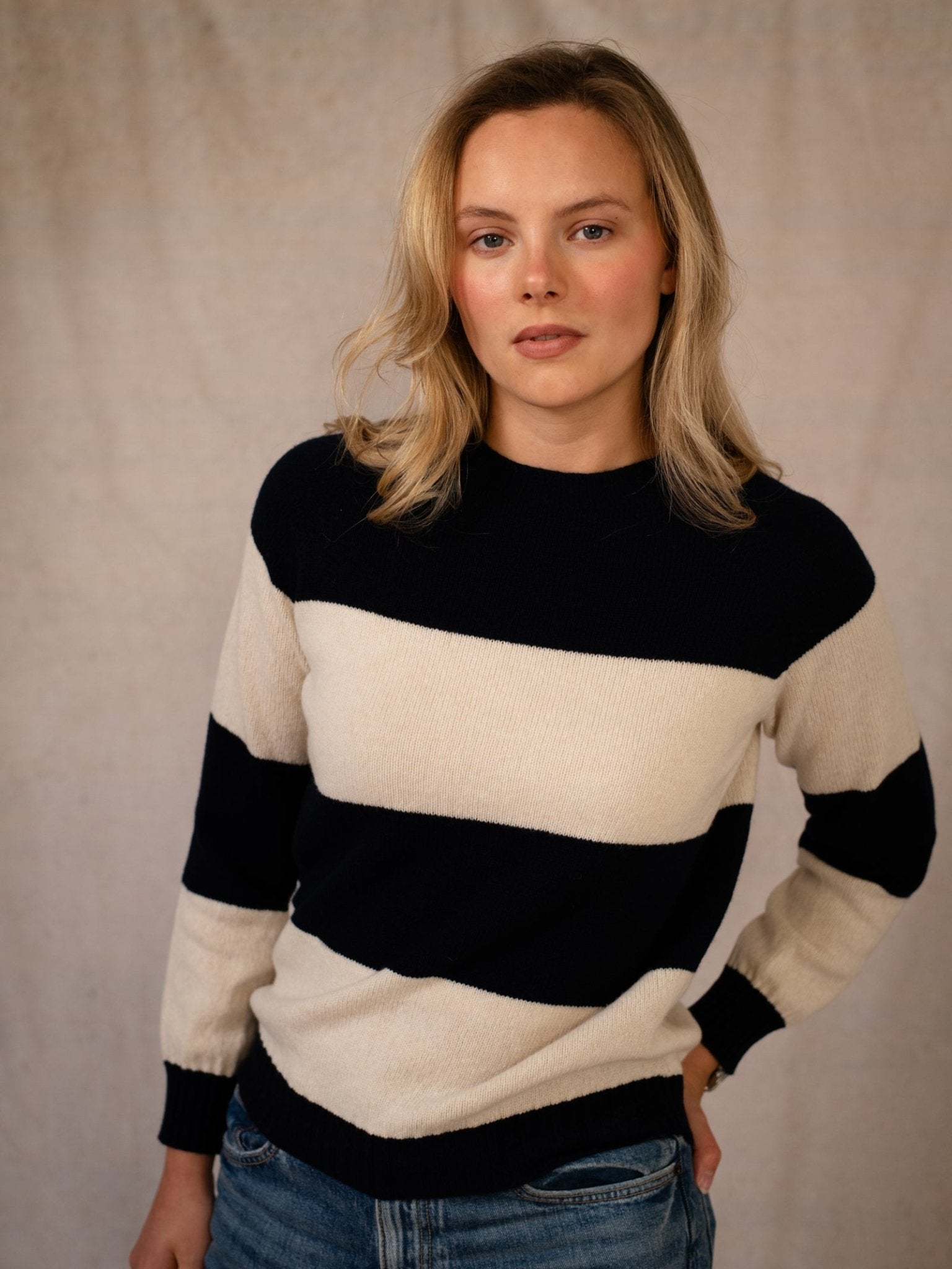 A woman with blonde hair wears the Geelong Stripe Crew by Campbell's of Beauly—a black and white striped jumper—paired with blue jeans. She stands before a beige background, looking calm with one hand on her hip.