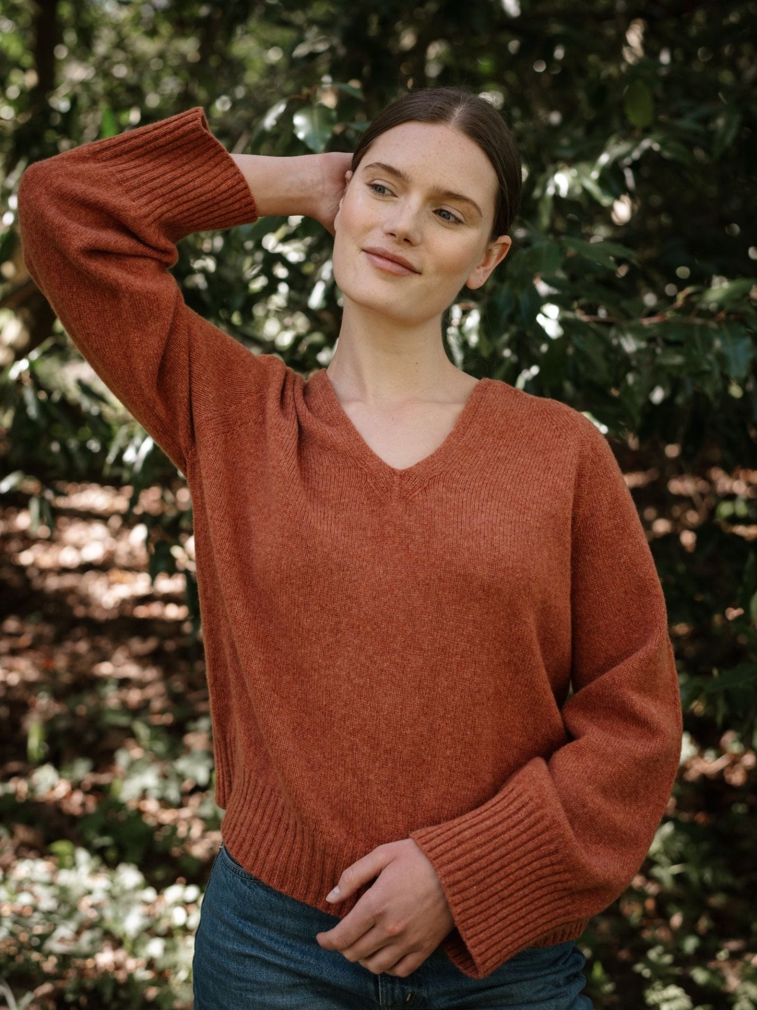 A woman in a rust Geelong Lambswool V Jumper by Campbell's of Beauly and blue jeans stands outdoors before green foliage, smiling gently with one hand behind her head.