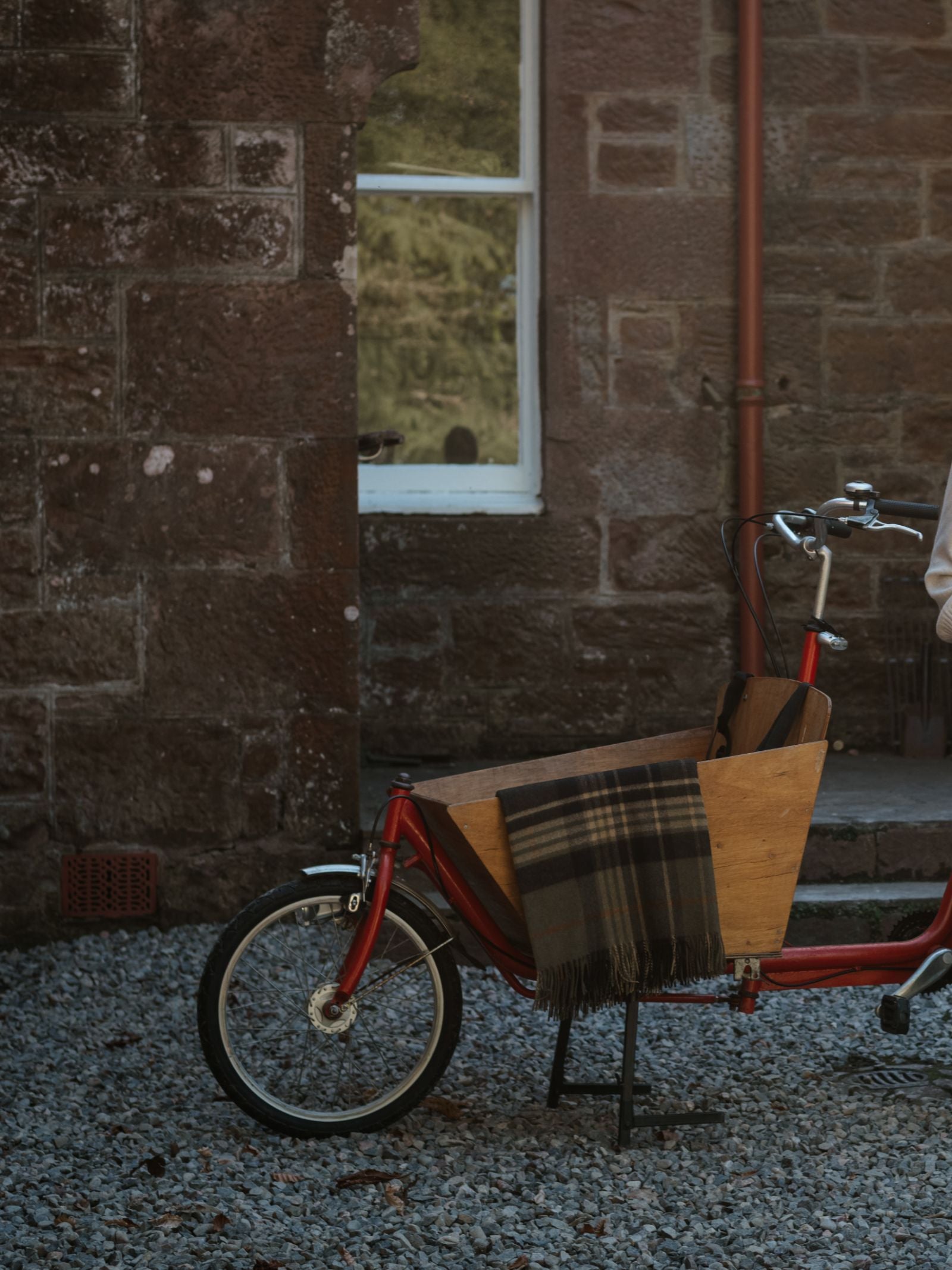 A red cargo bicycle with a wooden box is parked on gravel beside a brown stone building. Draped over the box is a cosy Campbell's of Beauly Glassburn Merino Throw.