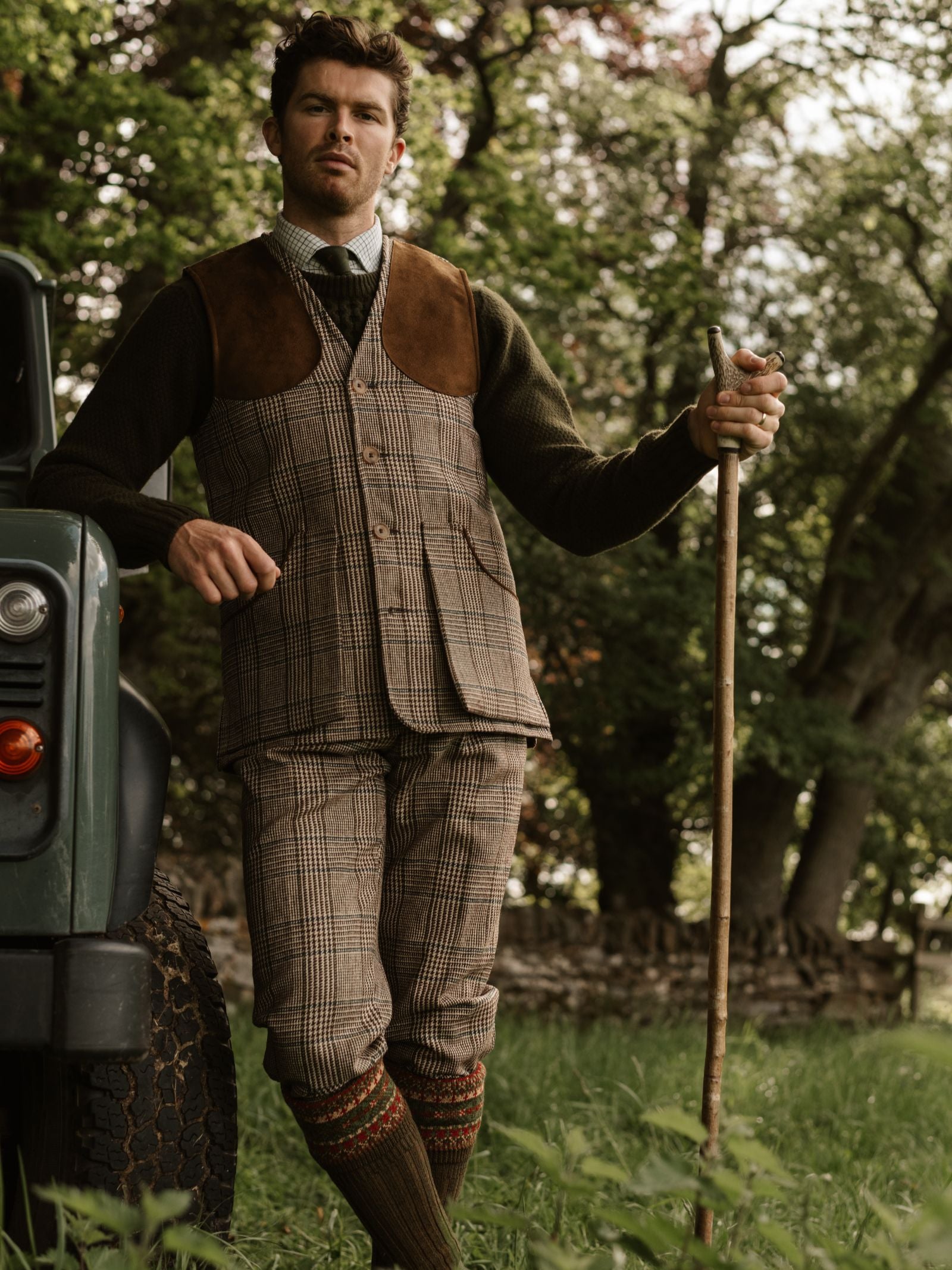 A man in country attire models Campbell's of Beauly Tweed Breeks, leaning on a green vehicle with a walking stick. He wears a Scottish tweed jacket, tie, and patterned socks, standing on grass with trees and a stone wall behind him.