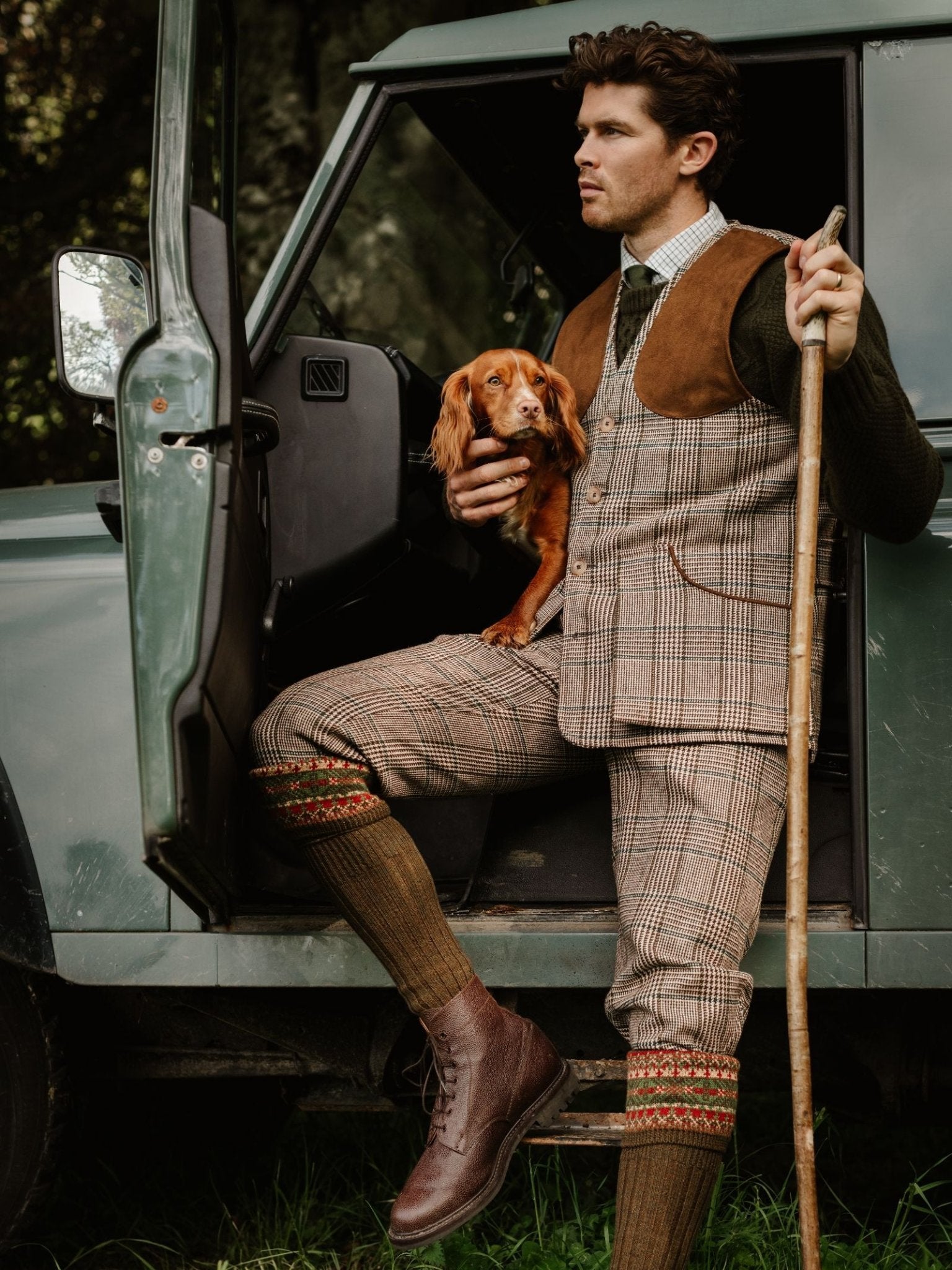 A man in Campbell's of Beauly Tweed Breeks sits in the open door of a green vehicle, holding a brown dog and walking stick, gazing into the distance with grass and trees in the background.