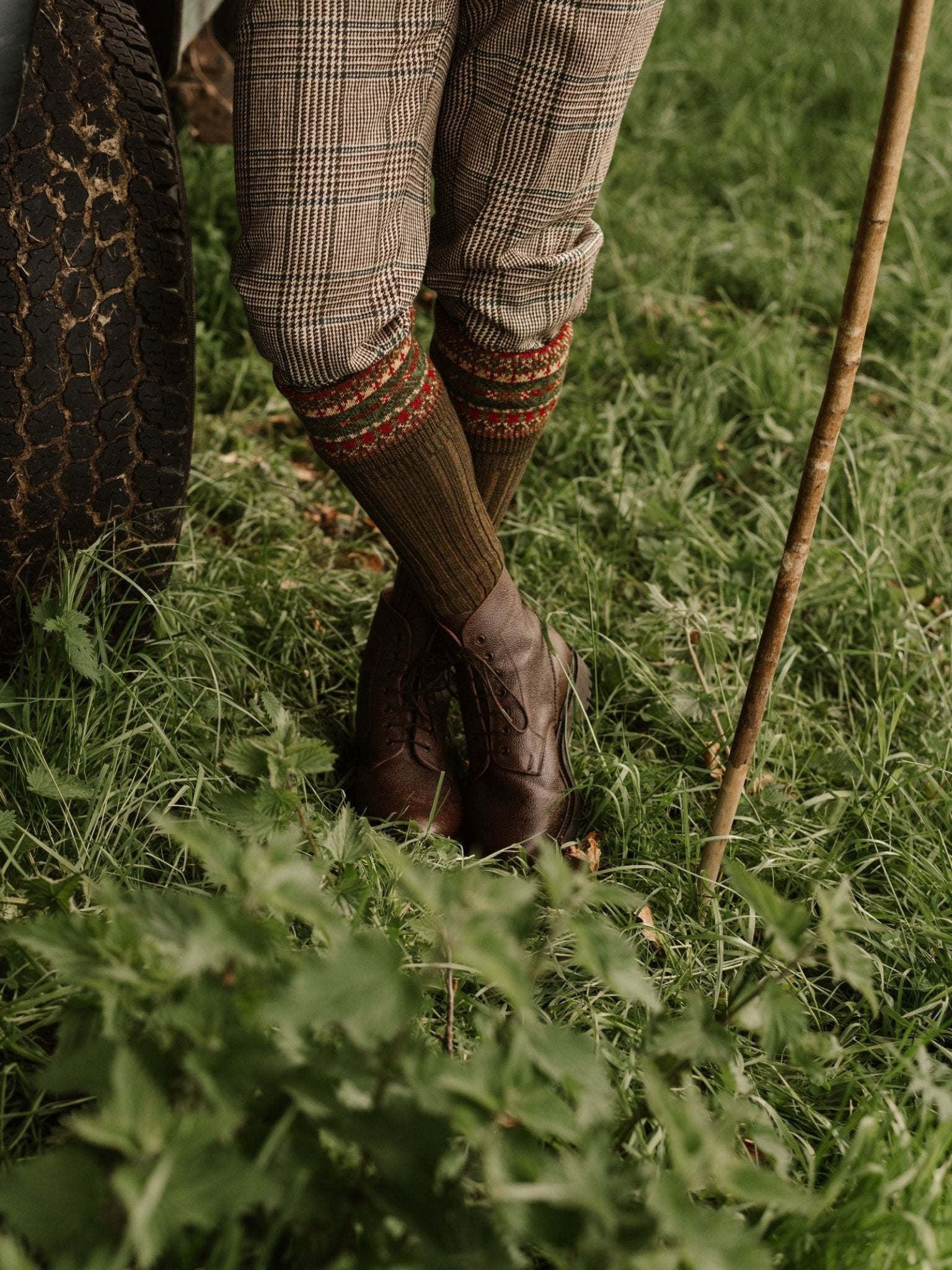 Wearing Campbell's of Beauly Tweed Breeks, classic country attire, brown boots, and patterned socks, a person stands on grass by a vehicle tire, holding a wooden walking stick.