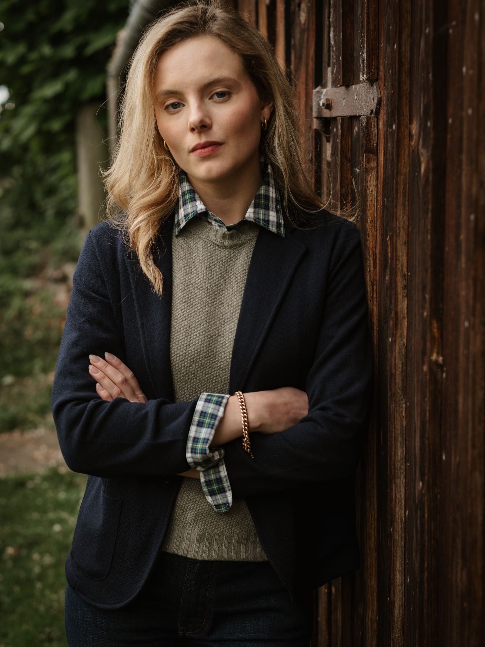 A blonde woman stands outdoors by a wooden wall, arms crossed, wearing a navy blazer, gray sweater, and the Campbell's of Beauly Cotton Tartan Shirt. She looks confidently at the camera with green foliage in the background.