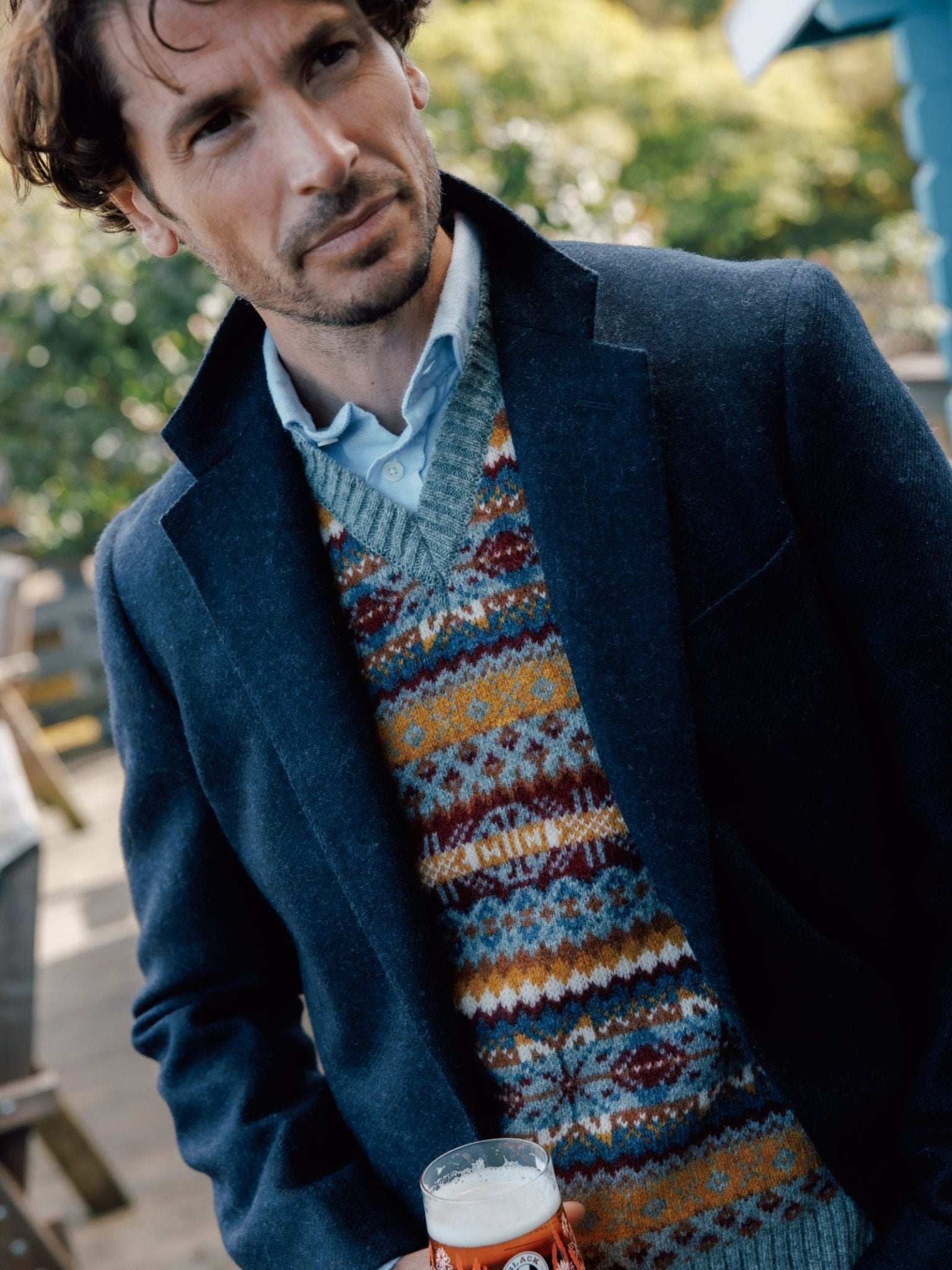 A man with wavy brown hair wears the Campbell's of Beauly Campbell's House Jacket over a blue shirt and patterned sweater vest, holding a beer outside with greenery and a blue building in the background.