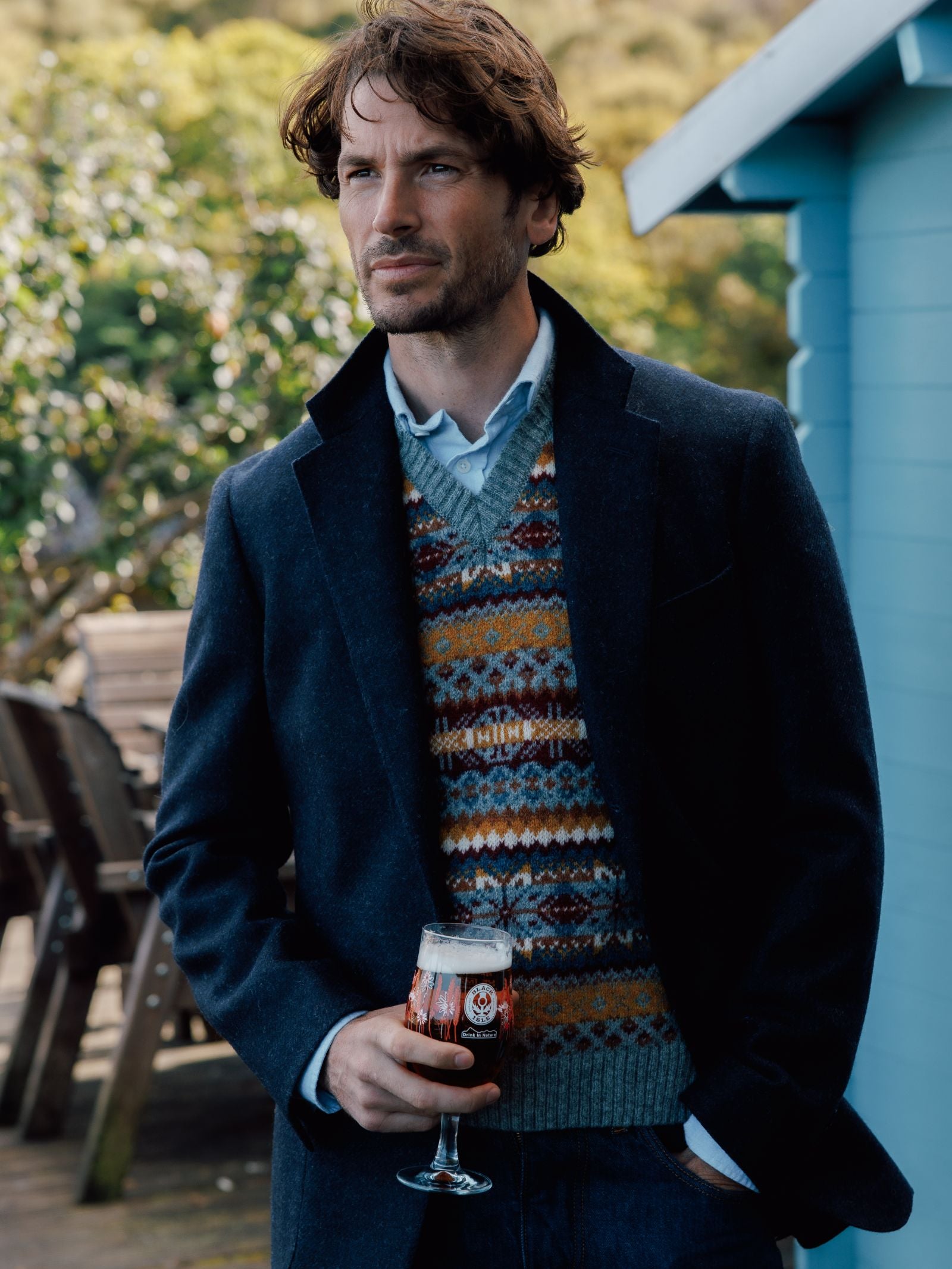 A man with wavy brown hair stands outdoors in a Campbell's House Jacket by Campbell's of Beauly over a colorful patterned sweater and blue shirt, holding a glass of dark beer with a blue building and greenery behind him.