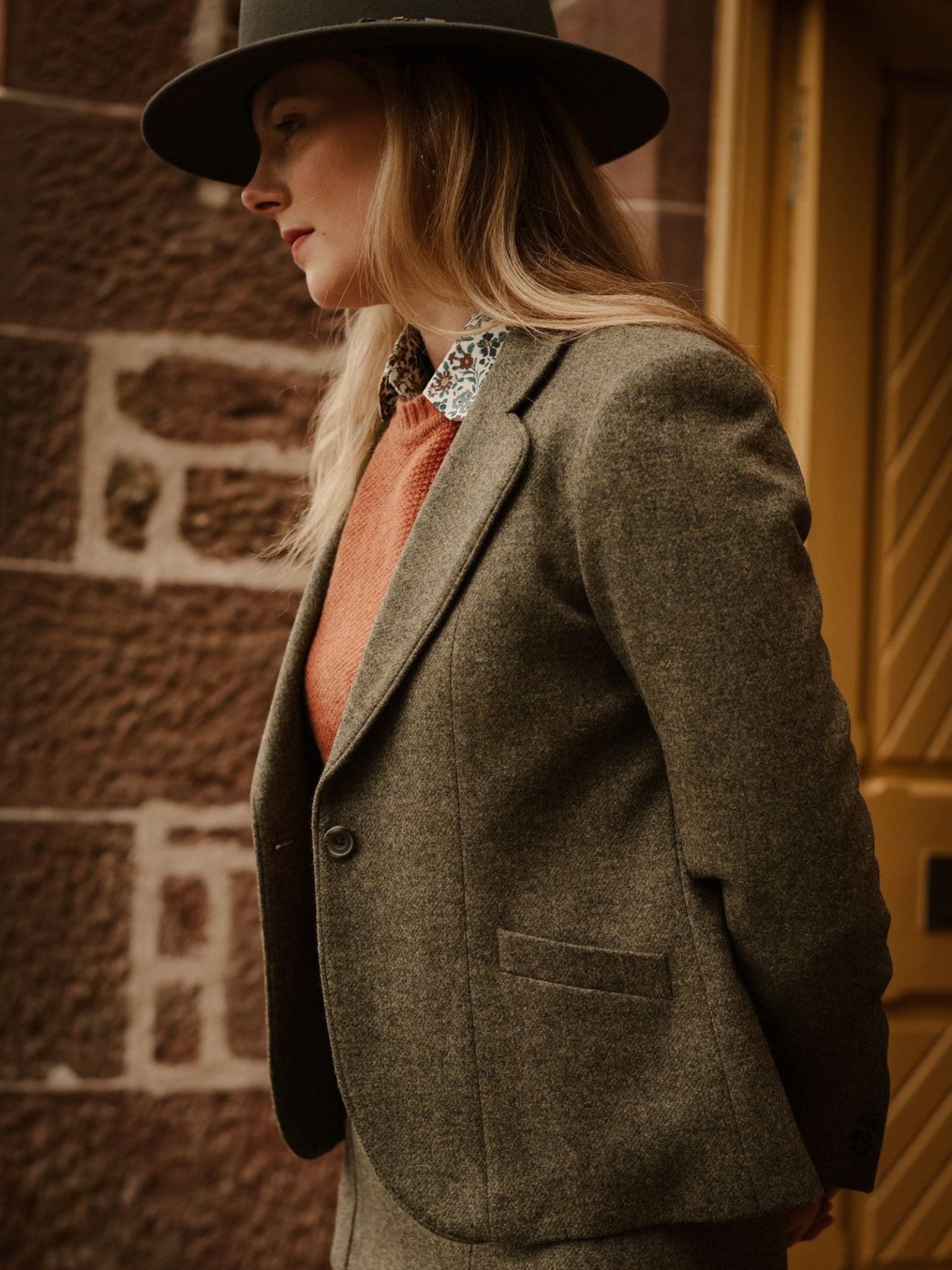 A person with long blonde hair, in a black hat and Campbell's of Beauly House Jacket, stands before a brown stone wall and yellow door, looking to the side.