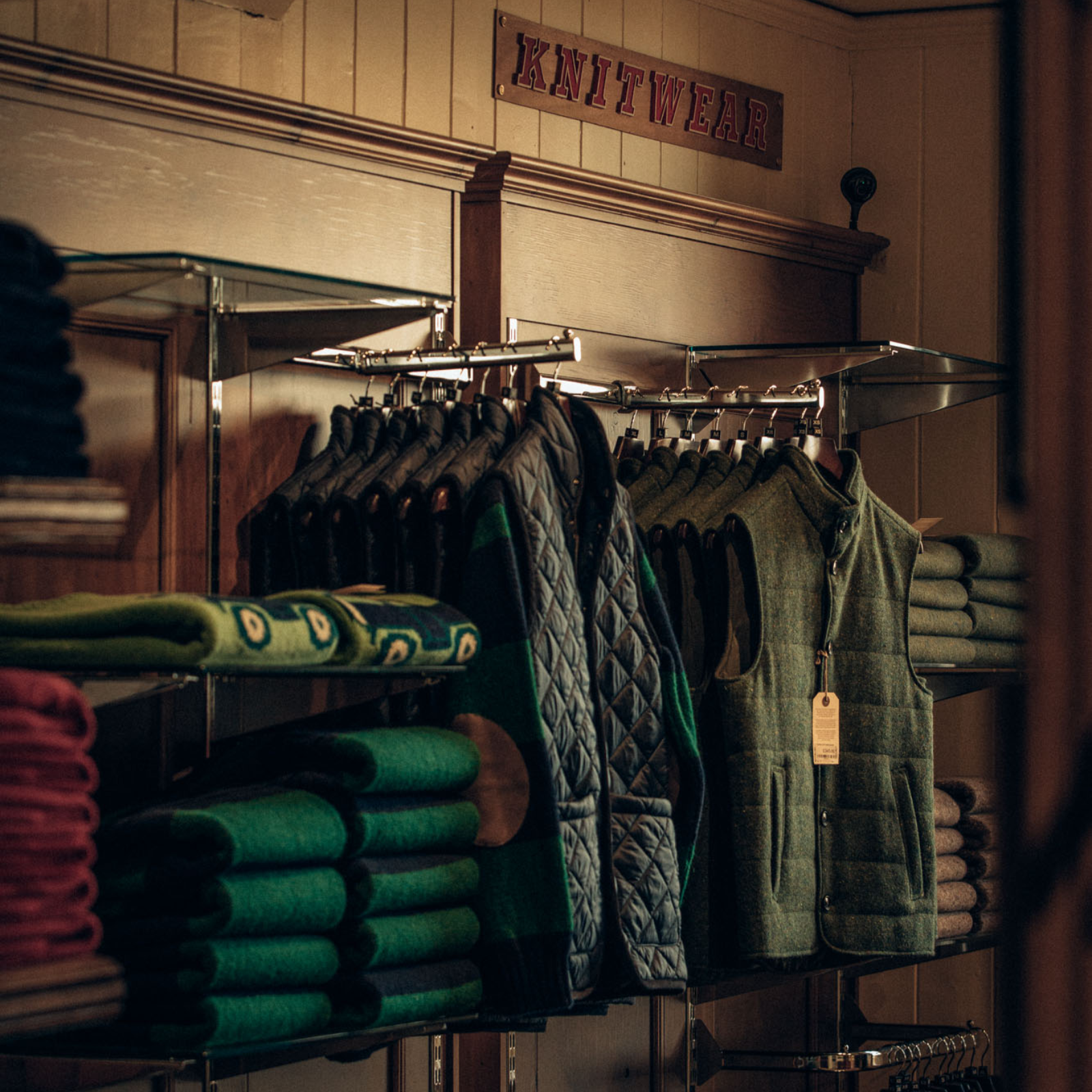 A cozy shop display features green and dark quilted vests on hangers, stacked green and burgundy sweaters, and a red Knitwear sign above wooden shelves. Warm lighting gives the scene a rustic feel.