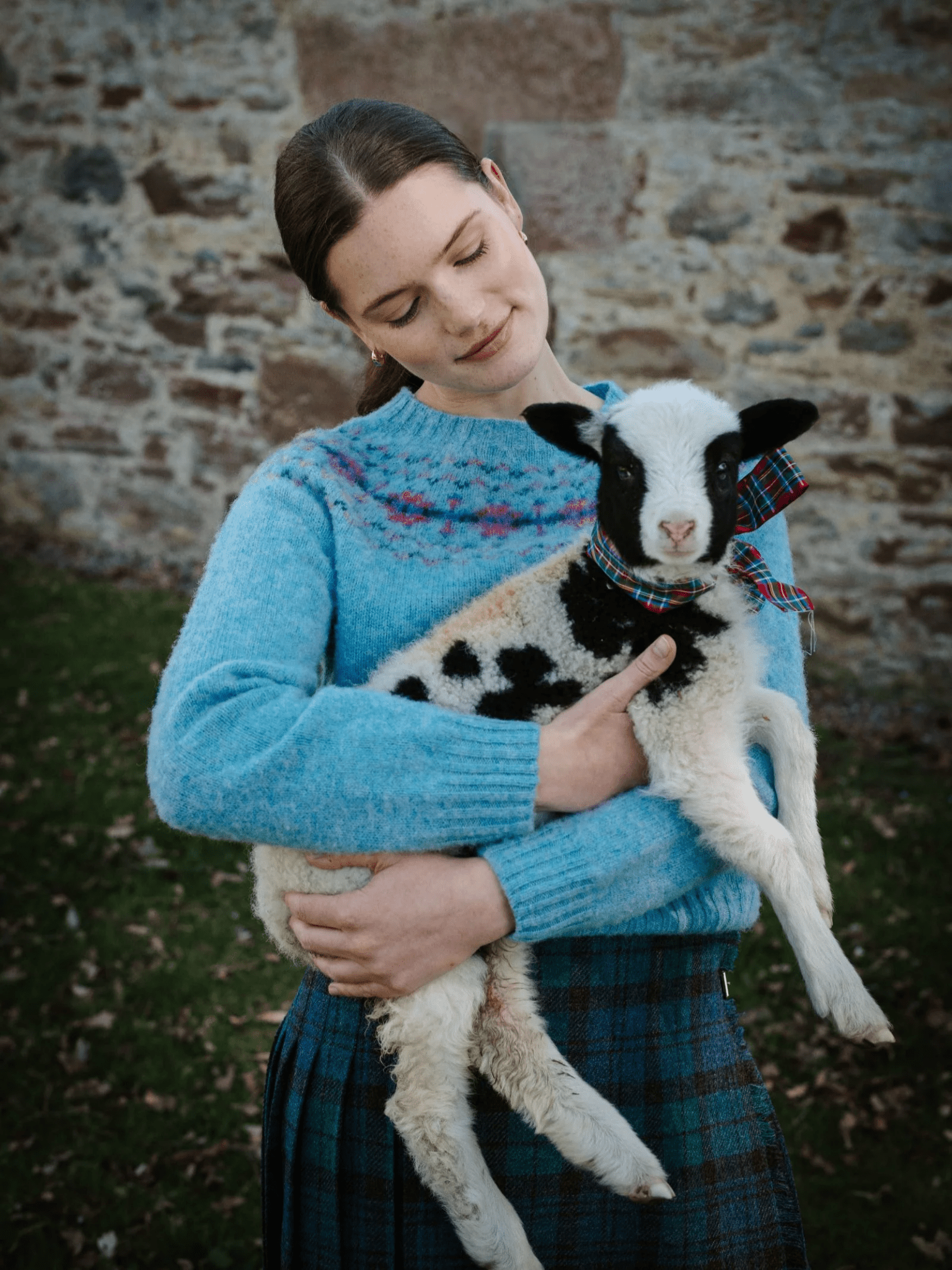 A woman in a Campbell's of Beauly Shaggy Cropped Fairisle Crew Jumper in Dewdrop and a plaid skirt gently holds a small black and white lamb with a tartan ribbon, standing outside by a stone wall on grass.
