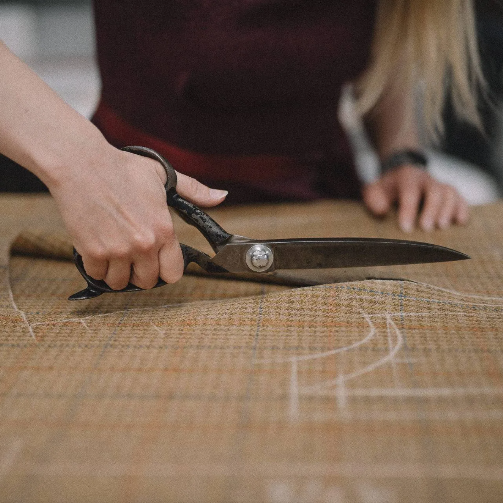 A person using large metal scissors to cut patterned fabric, with chalk markings visible on the cloth, suggesting tailoring or sewing work in progress.