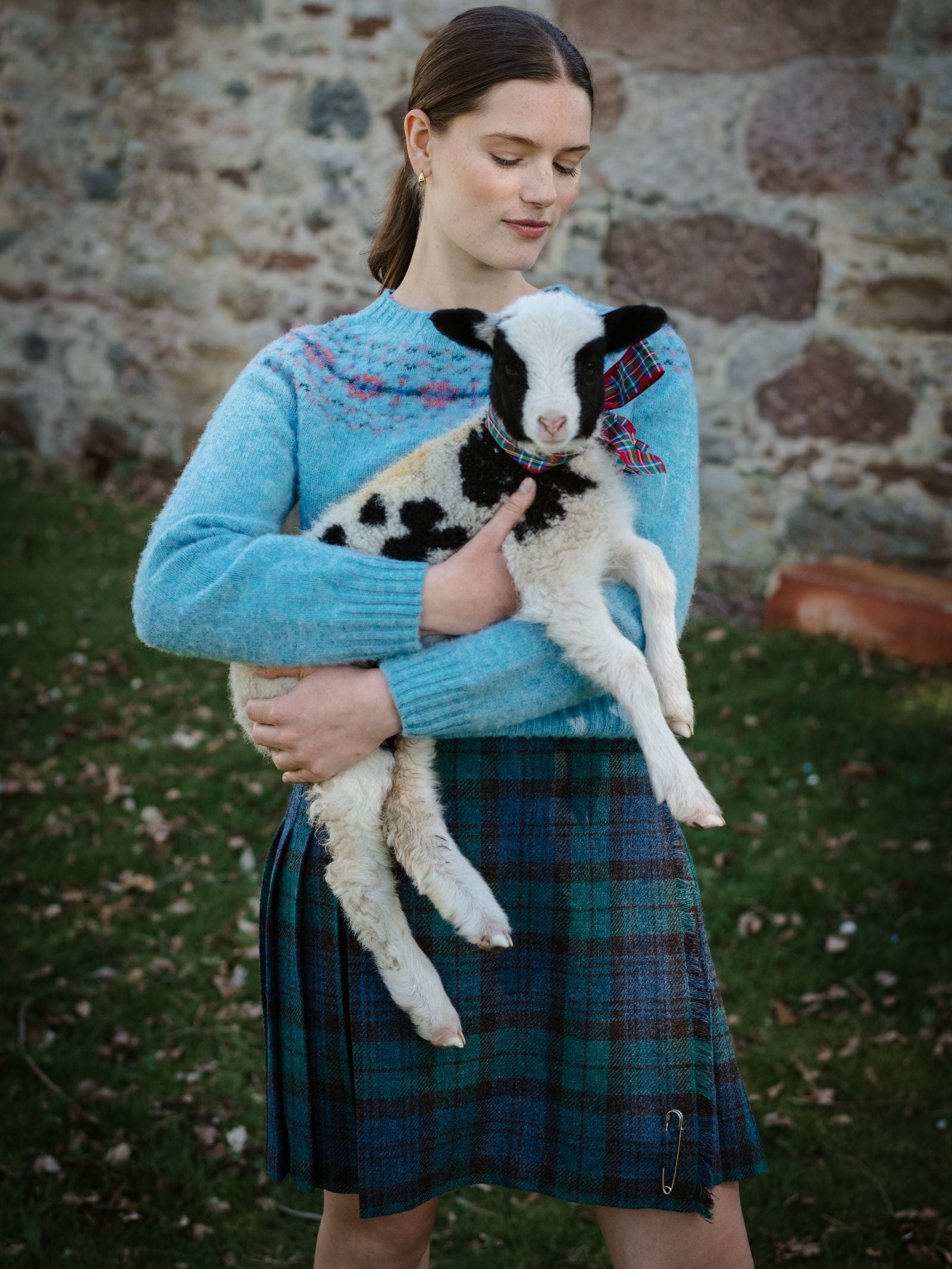 A woman wearing a Campbell's of Beauly Tweed Kilt stands outdoors holding a black-and-white lamb with a colorful plaid ribbon around its neck. A stone wall and grassy area are visible in the background.