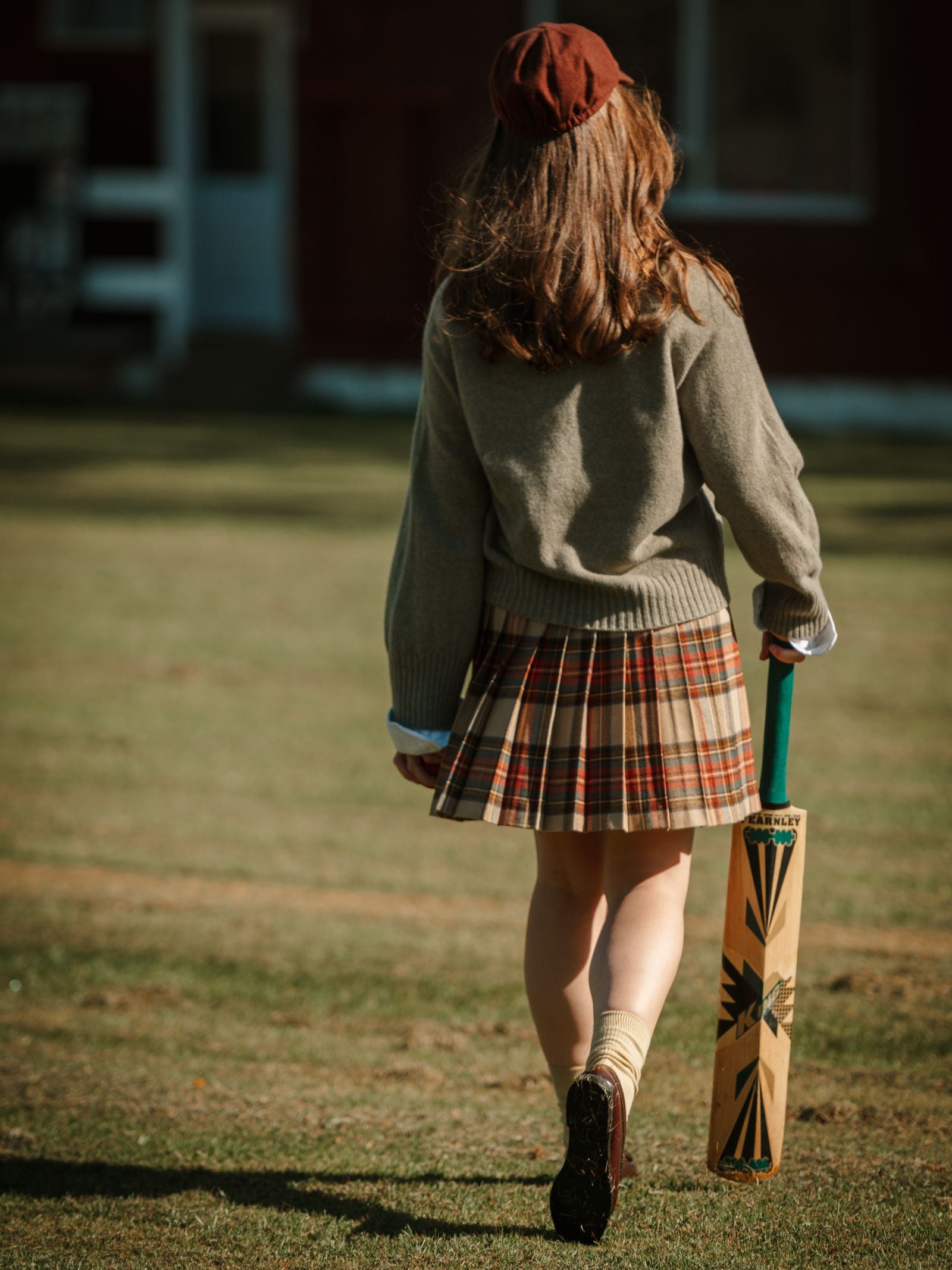 A girl wearing a Campbell's of Beauly Tweed Kilt with a sweater and cap walks on grass, holding a cricket bat at her side. Her back faces the camera, with a red building in the background.