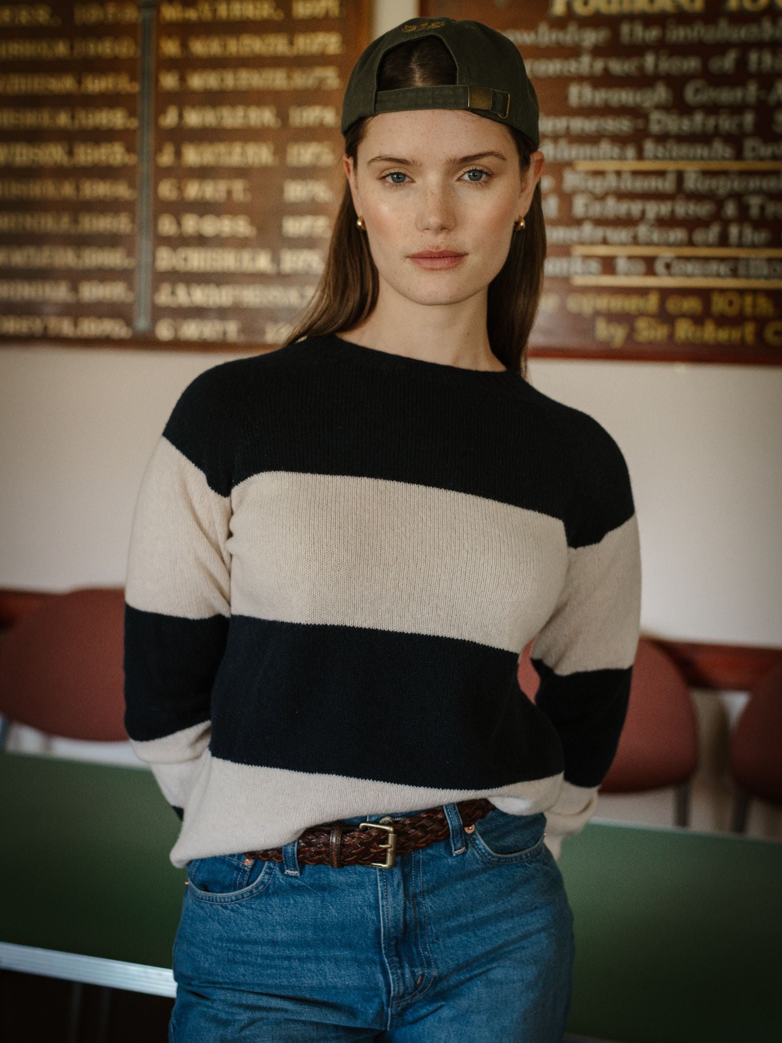 A woman indoors wears a green baseball cap backwards, blue jeans, and the Campbell's of Beauly Geelong Stripe Crew. Red chairs and a wooden board with text are visible in the background.