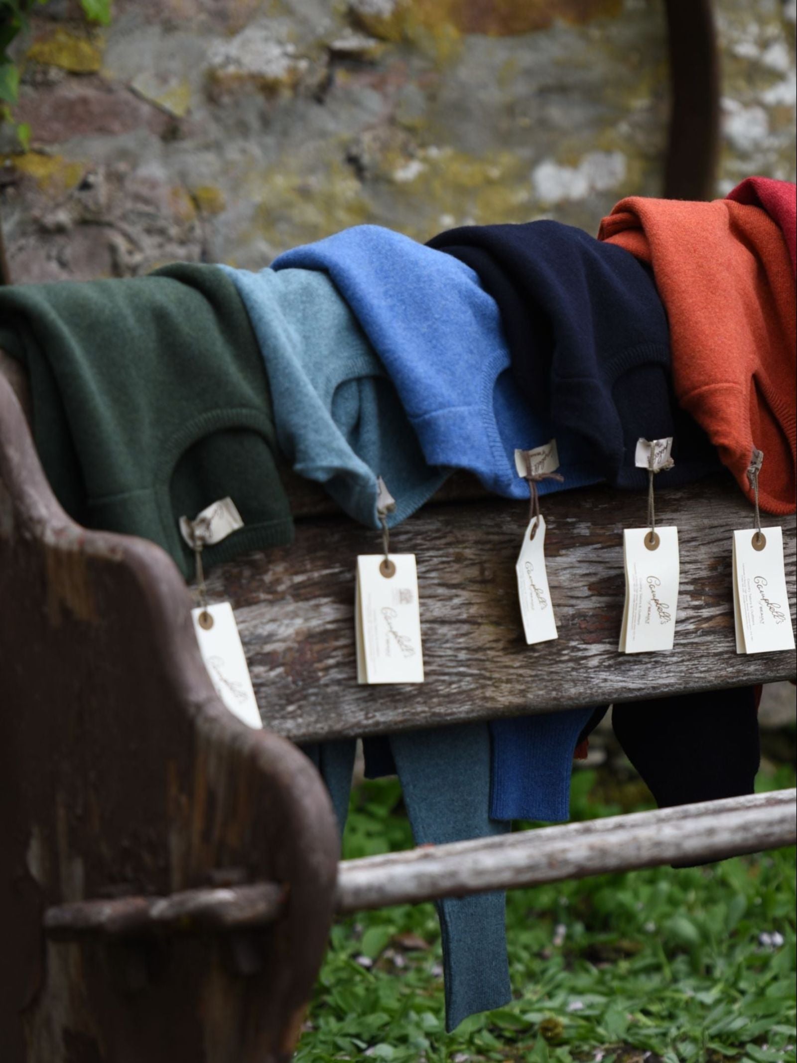 Five Campbell's of Beauly Lambswool Crew Neck Jumpers in green, blue, light blue, navy, and orange are neatly folded on a rustic wooden bench outdoors, each with a white tag. A stone wall and greenery complete the scene.