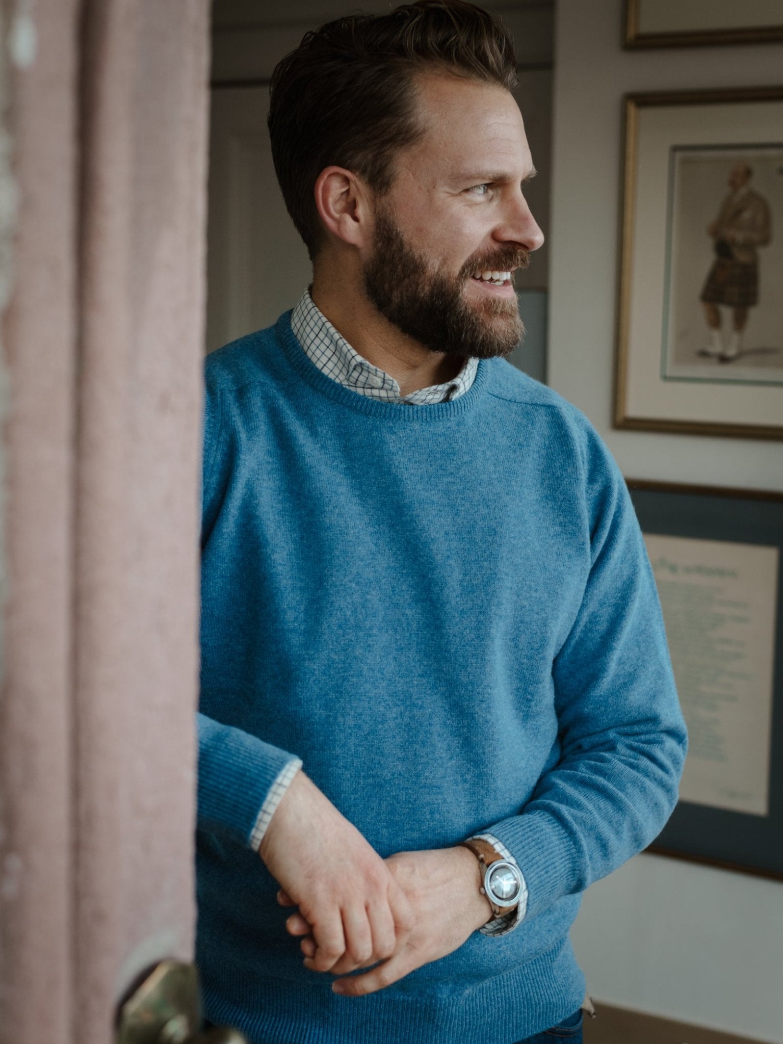 A bearded man stands indoors by a door, smiling to his left, wearing a Campbell's of Beauly Lambswool Crew Neck Jumper over a checkered shirt. Framed artwork and documents hang on the wall behind him.