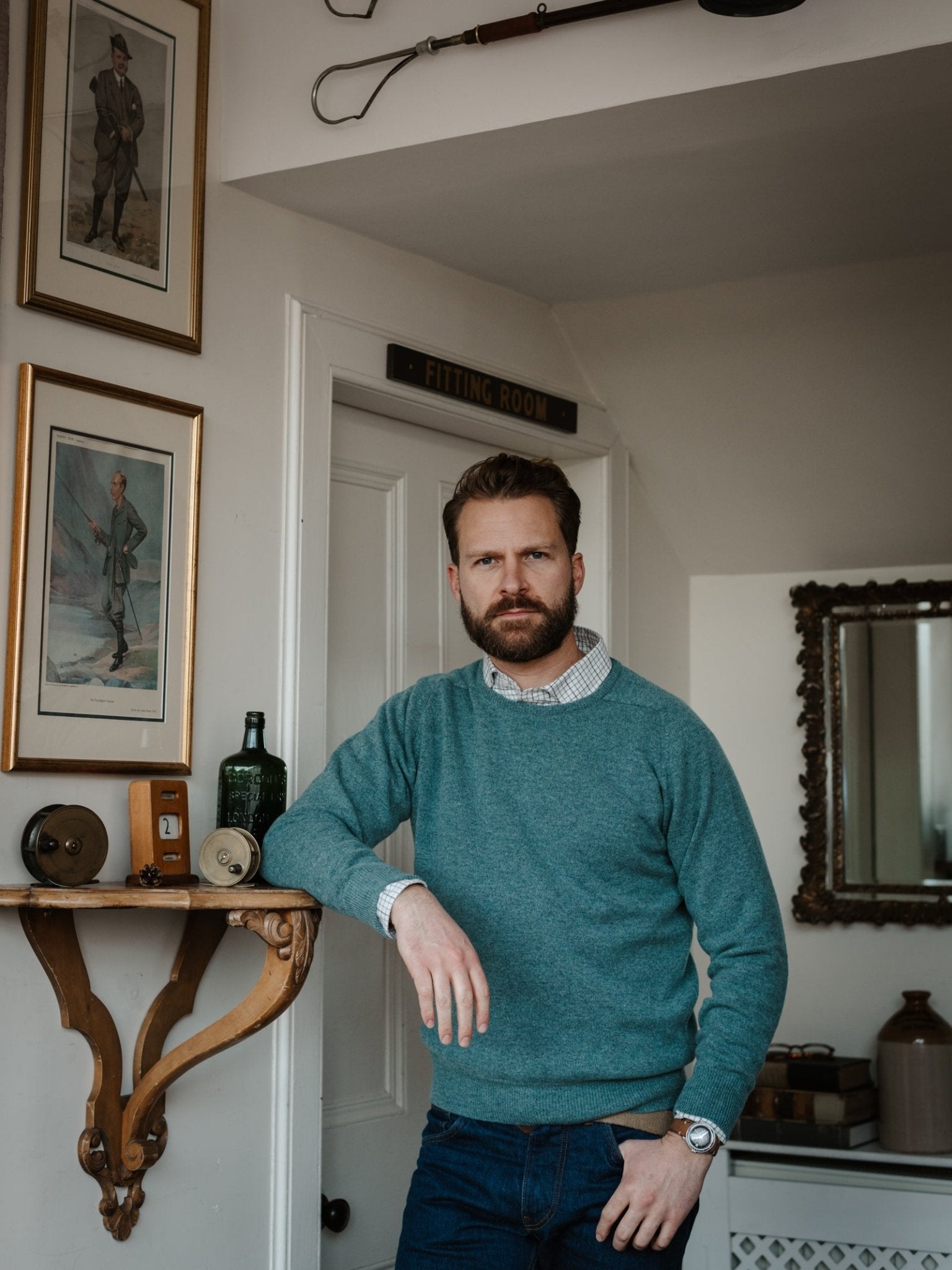 A bearded man in a Campbell's of Beauly Lambswool Crew Neck Jumper leans on a wooden shelf in a vintage room with framed pictures, fishing rods, and classic decor, looking at the camera with a neutral expression.