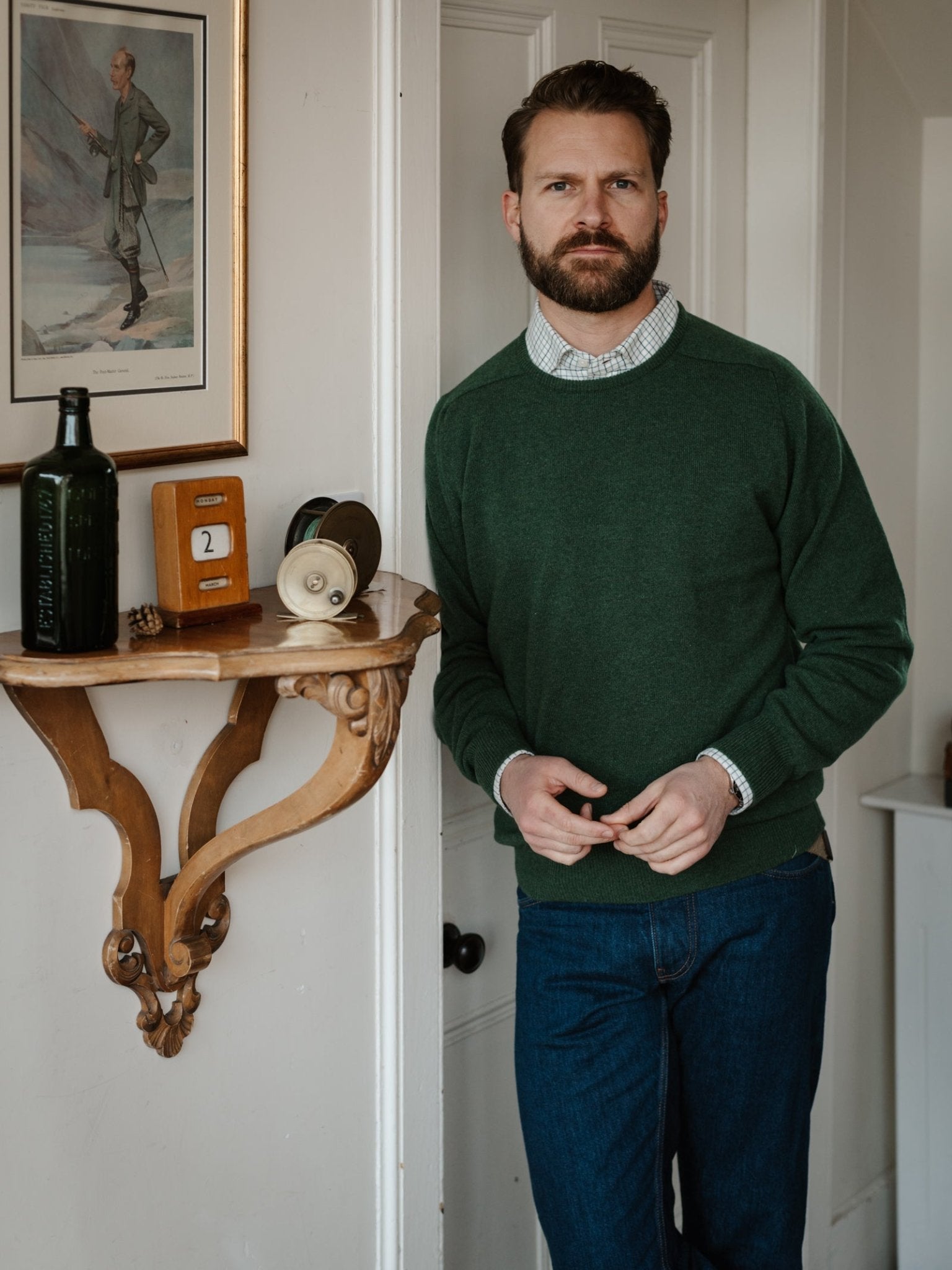 A bearded man in jeans stands indoors beside a wooden shelf with vintage items, wearing a green Lambswool Crew Neck Jumper by Campbell's of Beauly; above him hangs a framed painting.