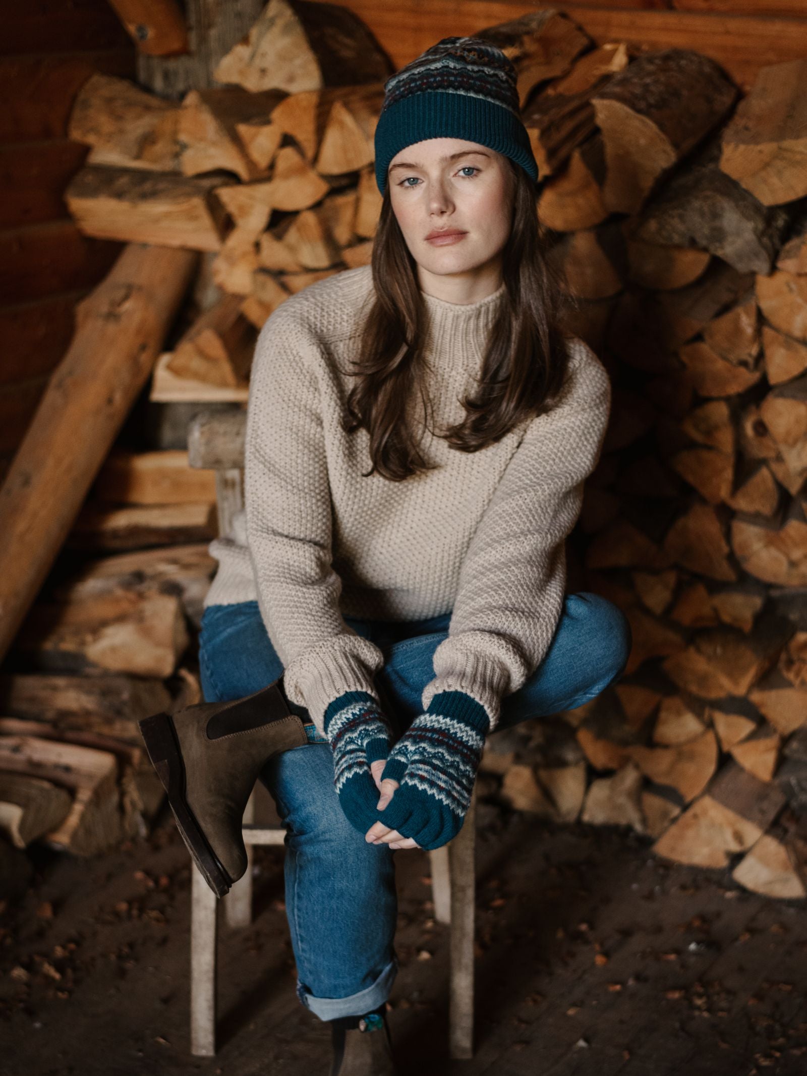 A woman in a beige sweater, blue jeans, and the Campbell's of Beauly Lambswool Fairisle Hat—this season’s winter staple—sits by stacked firewood in a rustic setting, gazing at the camera with a relaxed expression.