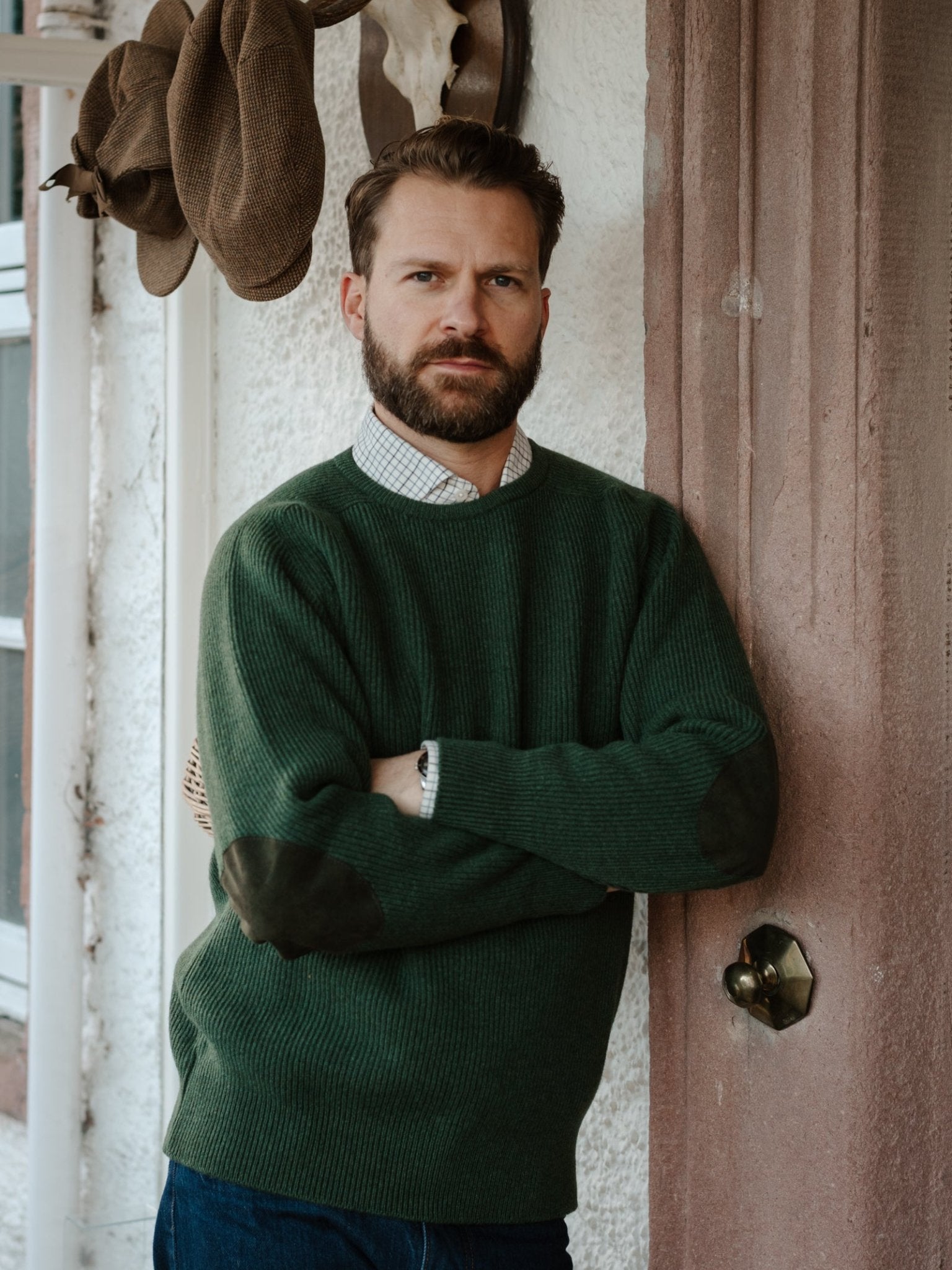 A bearded man stands with arms crossed against a pink doorway, wearing the Campbell's of Beauly Fisherman's Rib Crew Jumper in green over a checked shirt. Brown hats hang on the wall behind him.