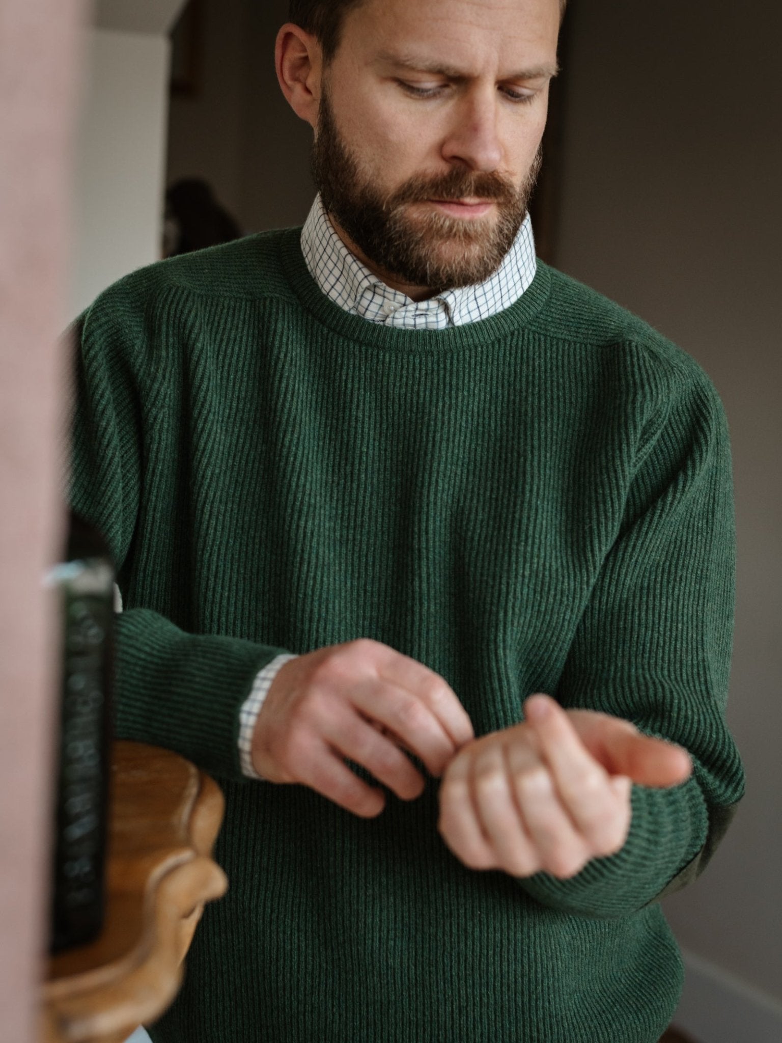 A bearded man, wearing the Campbell's of Beauly Fisherman's Rib Crew Jumper over a checkered shirt with suede elbow patches, looks down as he adjusts his cuff in a softly lit room.