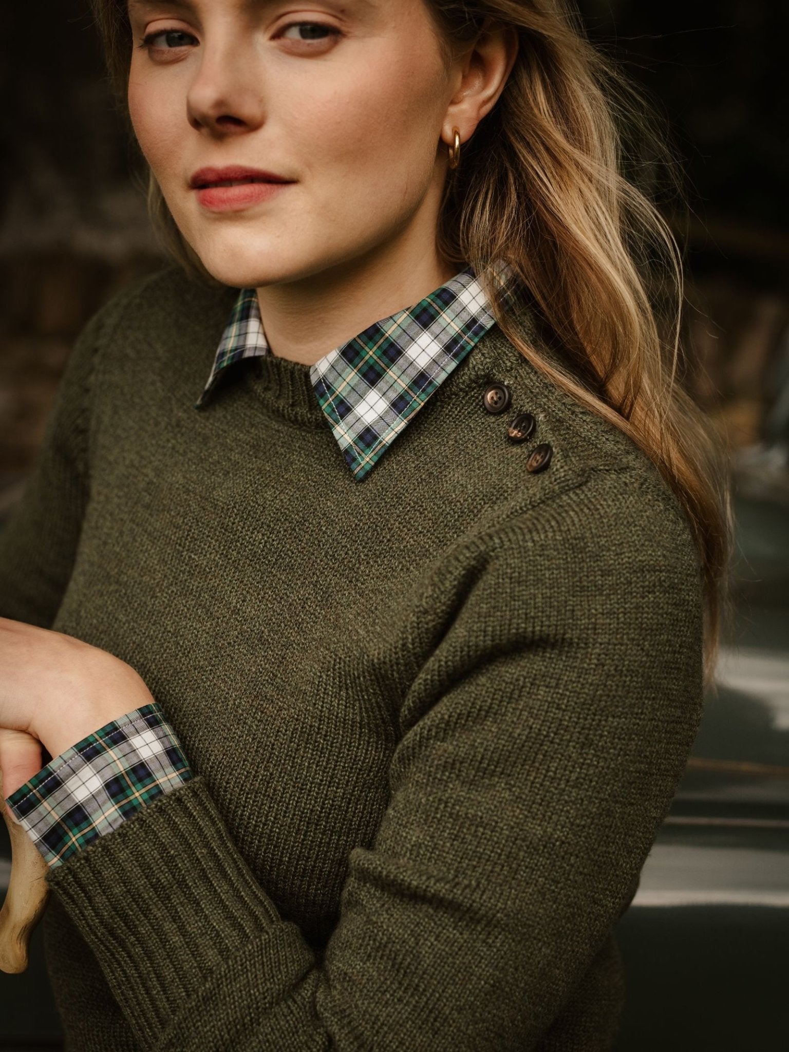 A woman stands outdoors looking to the side, wearing Campbell's of Beauly Lambswool Crew Neck Shooter Jumper in green with shoulder buttons, layered over a blue and green plaid collared shirt.