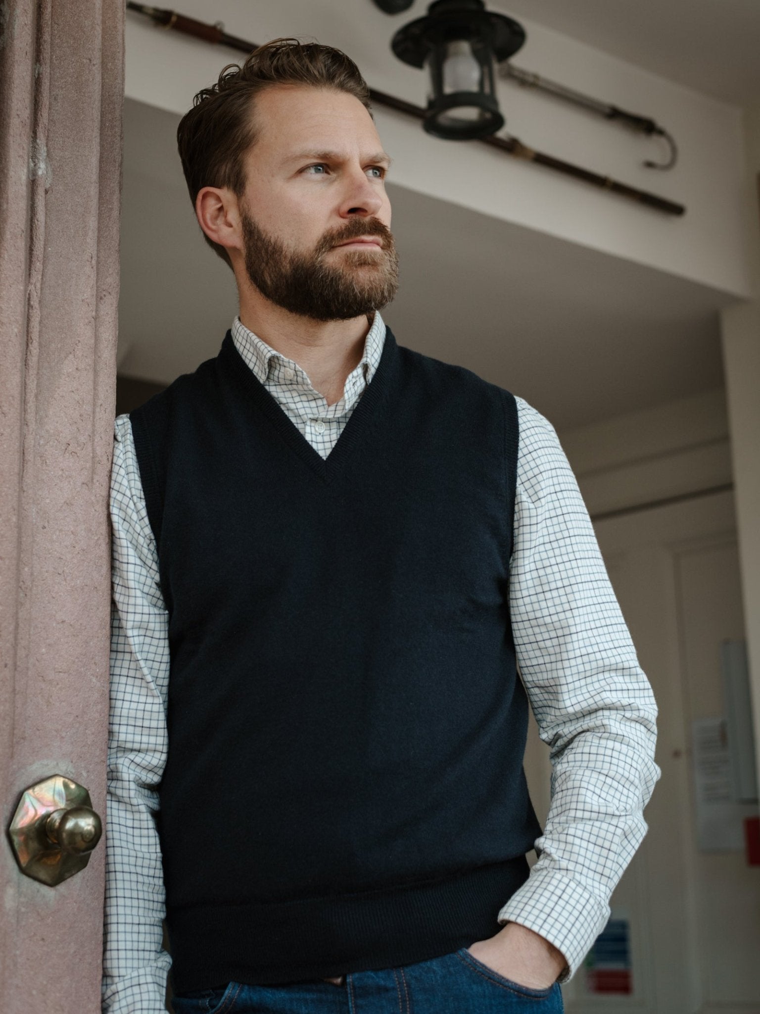 A bearded man in a checked shirt and Campbell’s of Beauly Auld Stock Lambswool V-Neck Slipover stands by a doorway, gazing thoughtfully into the distance indoors.