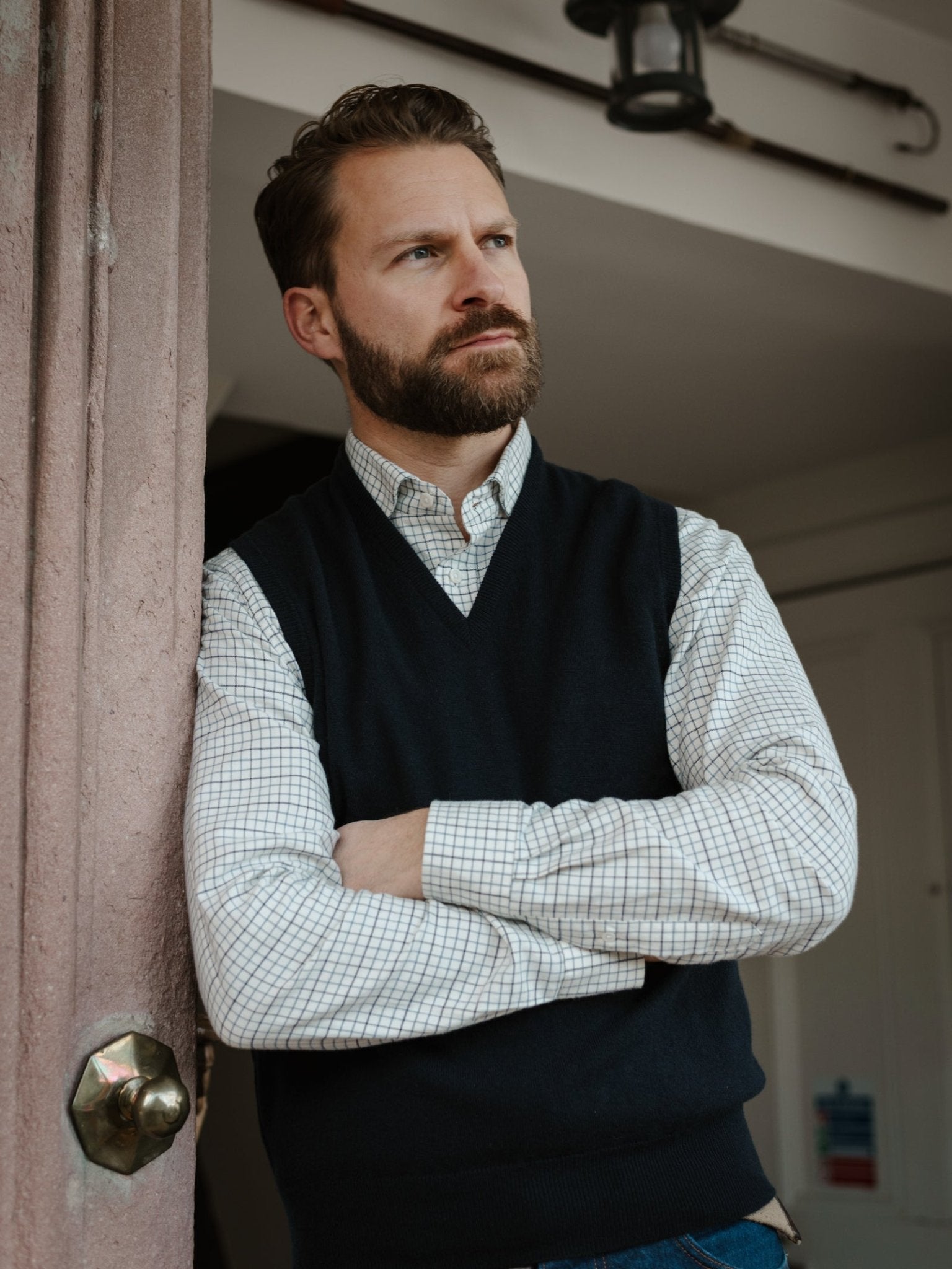 A bearded man stands indoors, leaning against a pinkish door frame with arms crossed. He wears a checkered shirt and the Campbell’s of Beauly Auld Stock Lambswool V-Neck Slipover, gazing thoughtfully into the distance.