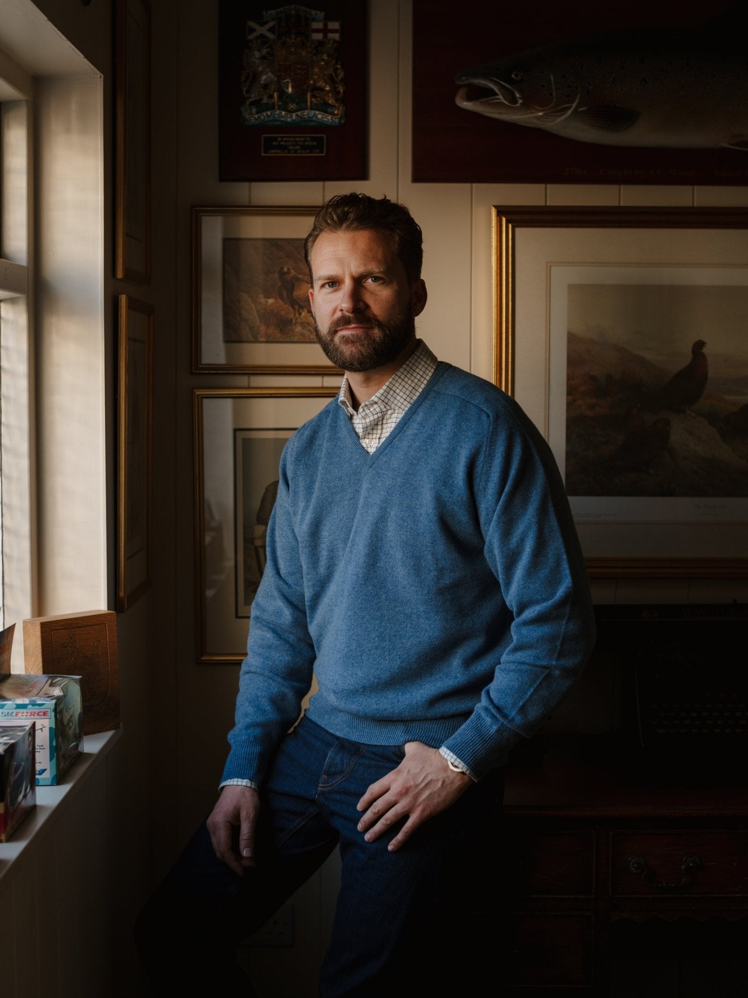 A bearded man with short hair sits on a windowsill, wearing the Campbell's of Beauly Lambswool V-Neck Jumper over a collared shirt and jeans, in a room adorned with framed art and a mounted fish.