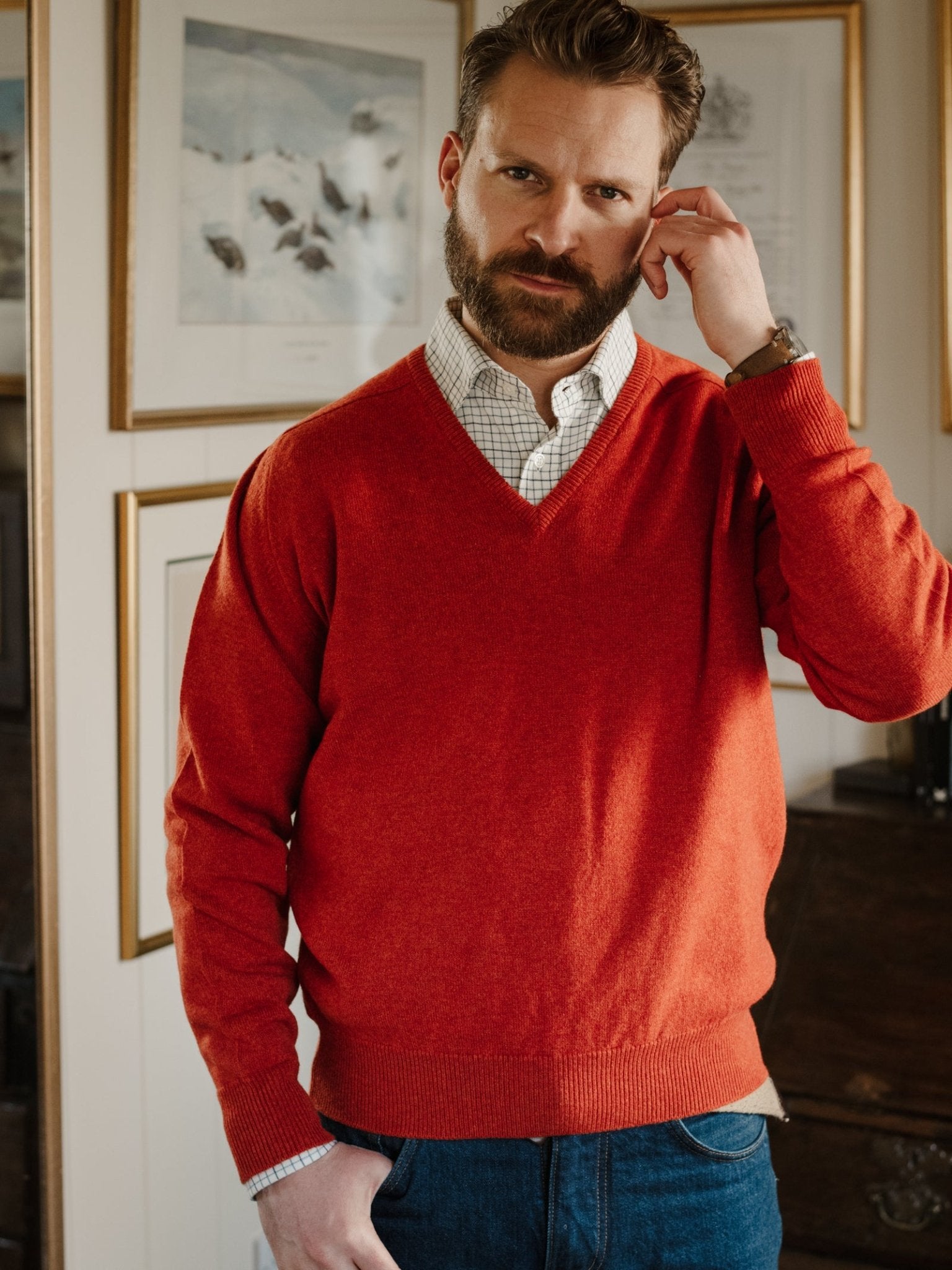 A bearded man wearing a red Lambswool V-Neck Jumper from Campbell's of Beauly over a collared shirt and blue jeans stands indoors, touching his ear. Framed art and a wooden cabinet appear in the background.