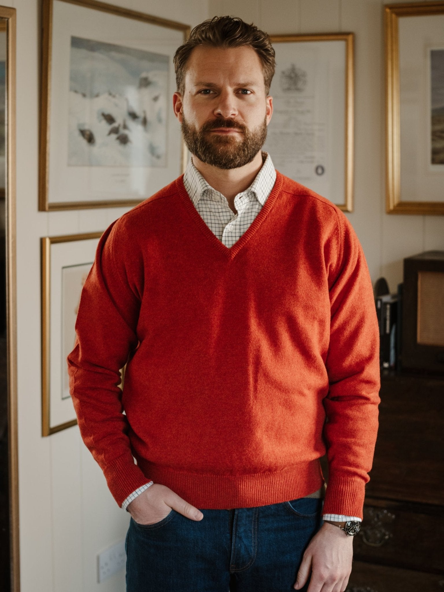 A bearded man stands indoors with one hand in his pocket, dressed in a Campbell's of Beauly Lambswool V-Neck Jumper over a check shirt and jeans. Framed artwork and certificates decorate the wall behind him.