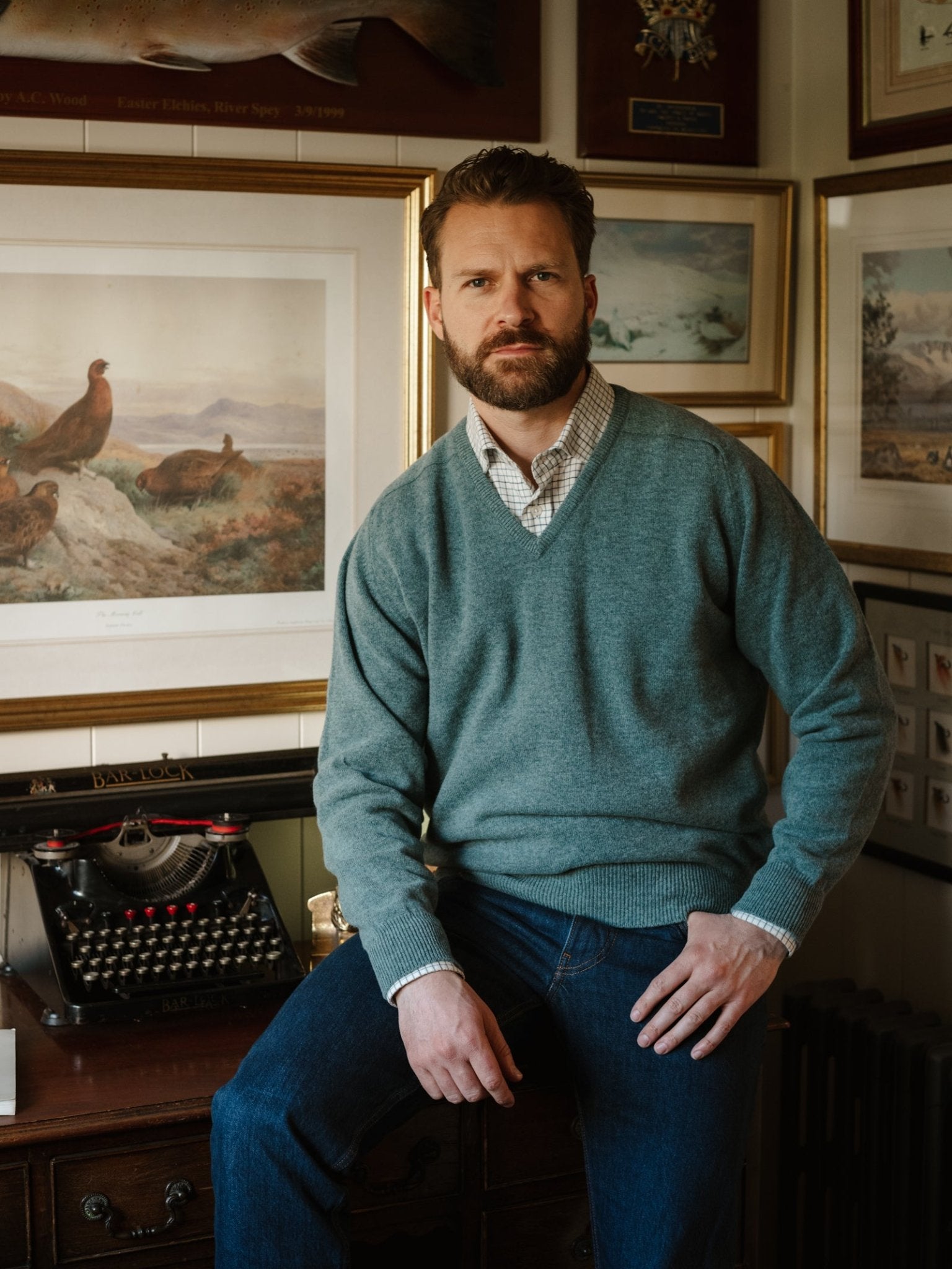 A bearded man sits at a desk, wearing the Campbell's of Beauly Lambswool V-Neck Jumper over a checked shirt and jeans. Behind him are framed art and a vintage typewriter, creating a cozy, vintage-inspired atmosphere.
