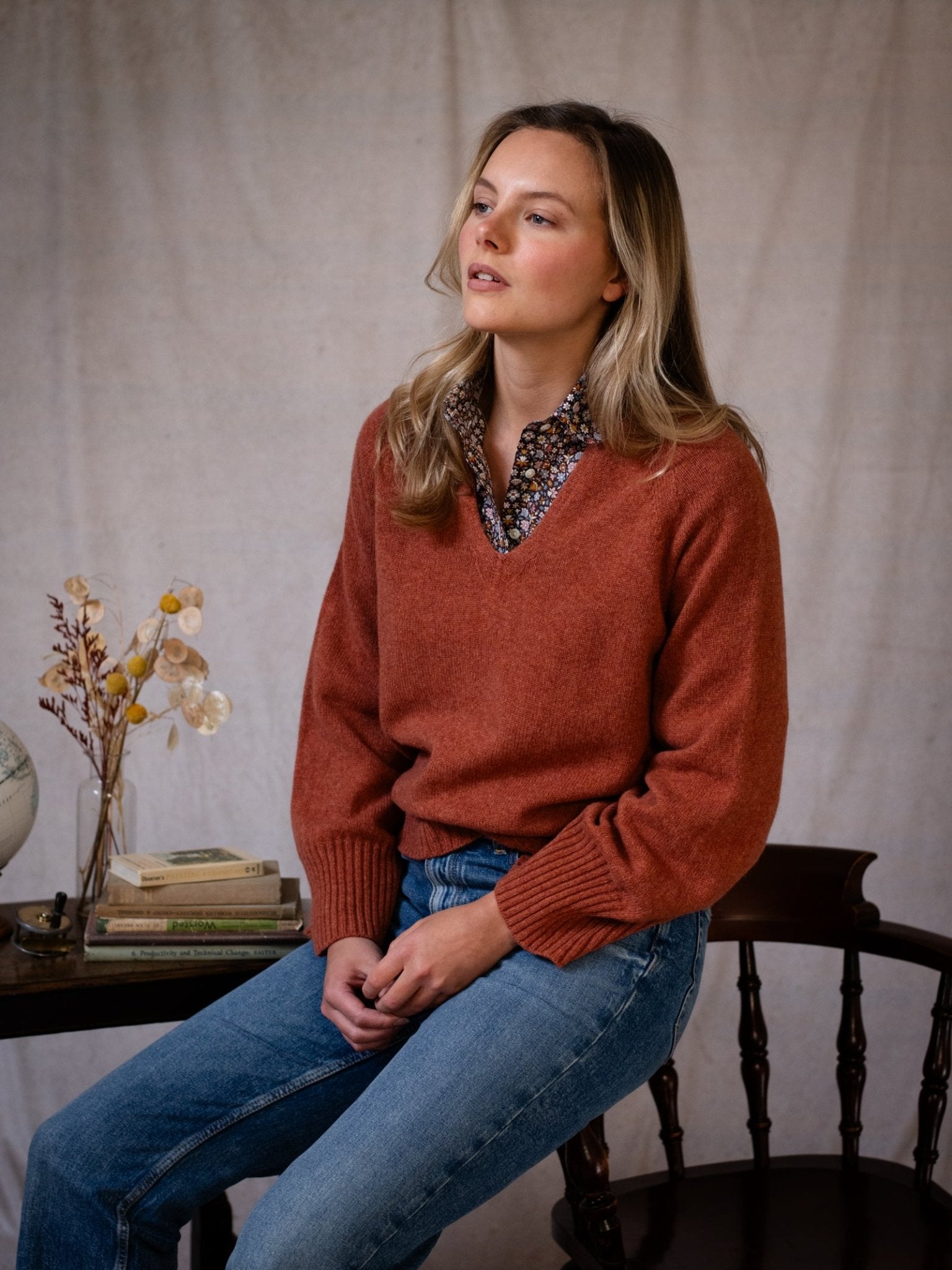 A woman with long blonde hair wears a rust-colored Campbell's of Beauly Geelong Lambswool V Jumper over a floral shirt and blue jeans. She sits on a wooden chair beside books, a globe, dried flowers, with neutral fabric as the backdrop.