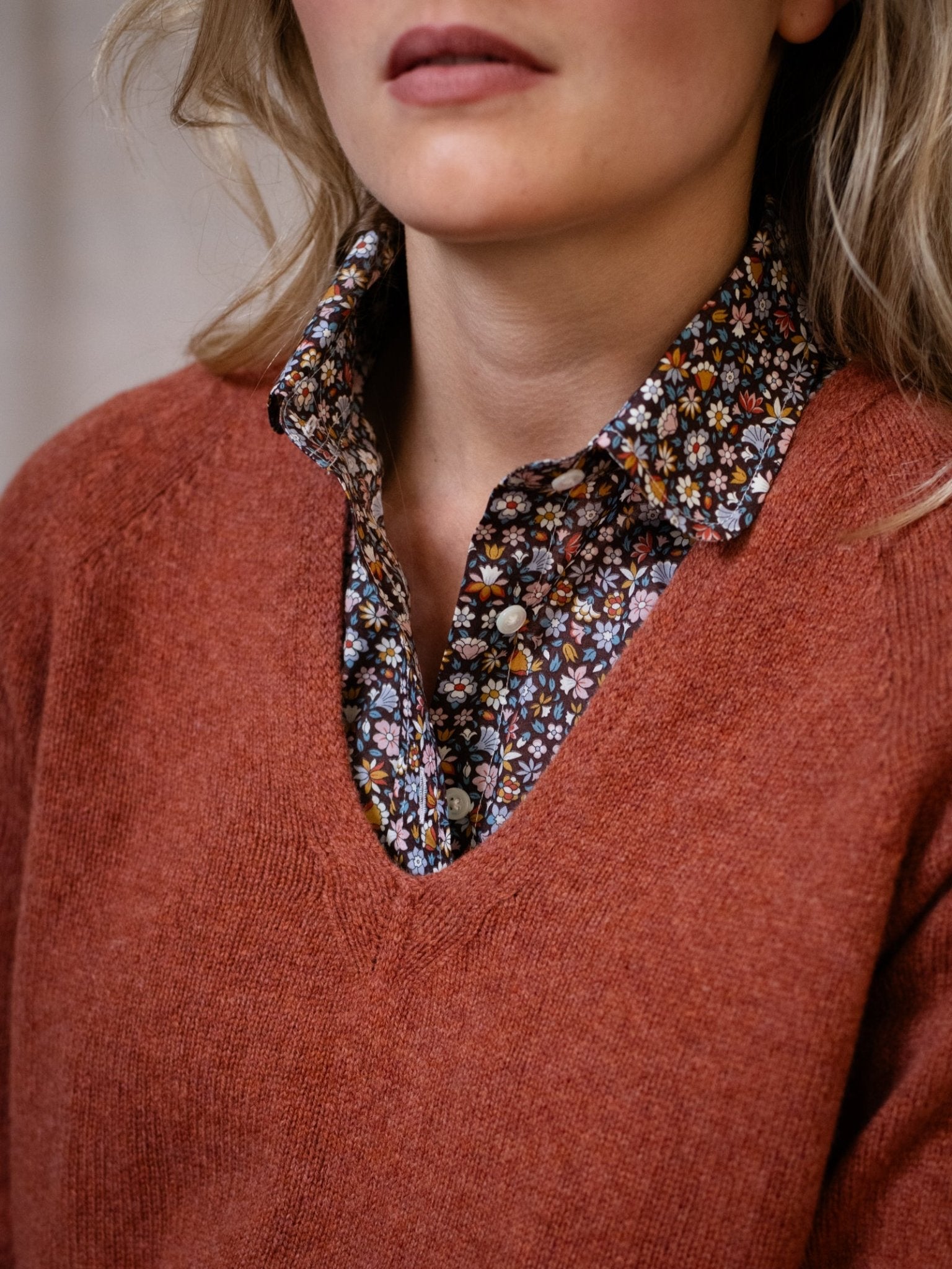 A woman with loose blonde hair, visible from her lower face to upper torso, wears a rust Geelong Lambswool V Jumper by Campbell's of Beauly over a black floral button-up shirt.