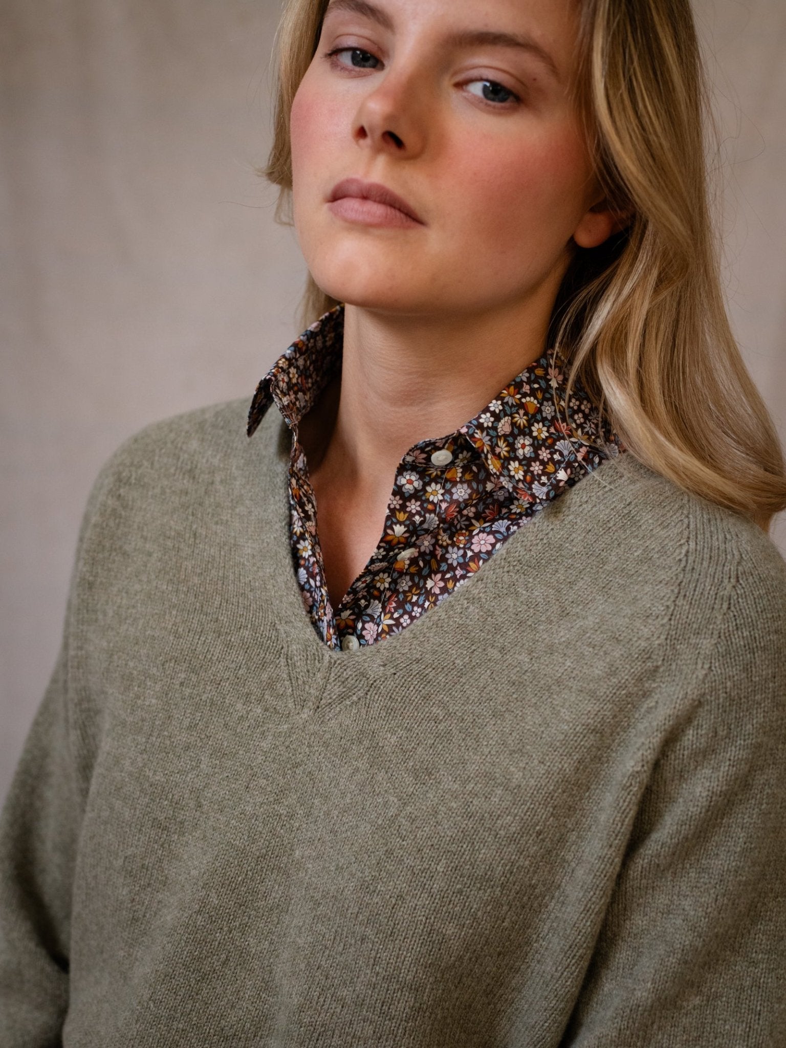 A person with long blonde hair wears a light olive-green Geelong Lambswool V Jumper by Campbell's of Beauly over a floral collared shirt, looking slightly away with a neutral expression against a plain, soft-toned background.