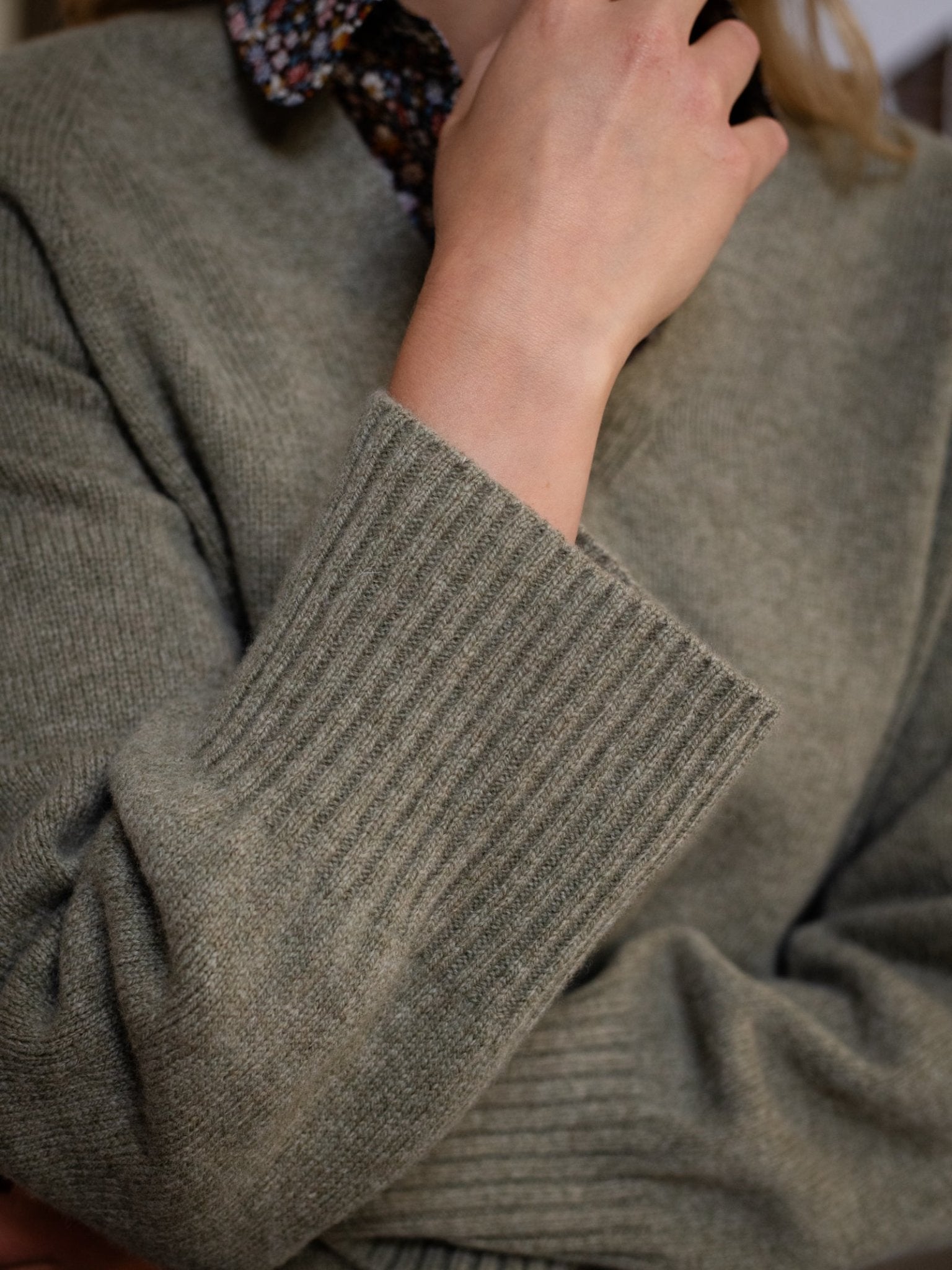 A person in a textured, ribbed gray-green Campbell's of Beauly Geelong Lambswool V Jumper with a floral-collared shirt holds their hand near their chin, partially obscuring their face.