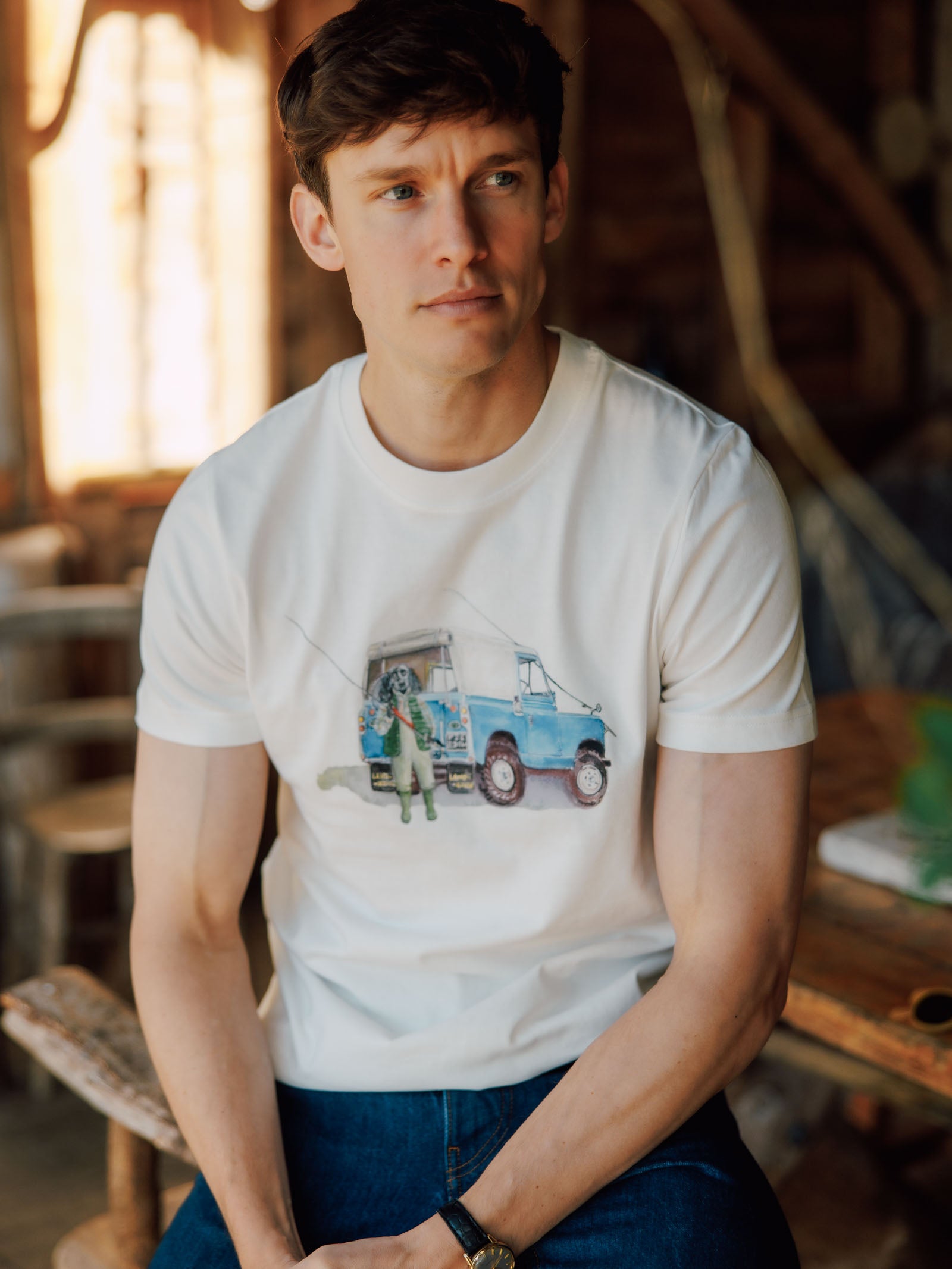 A young man with short brown hair sits indoors on a wooden chair, wearing the Campbell's of Beauly Land Rover Cocker T-Shirt, featuring a blue camper van and people print, capturing a relaxed classic country style.