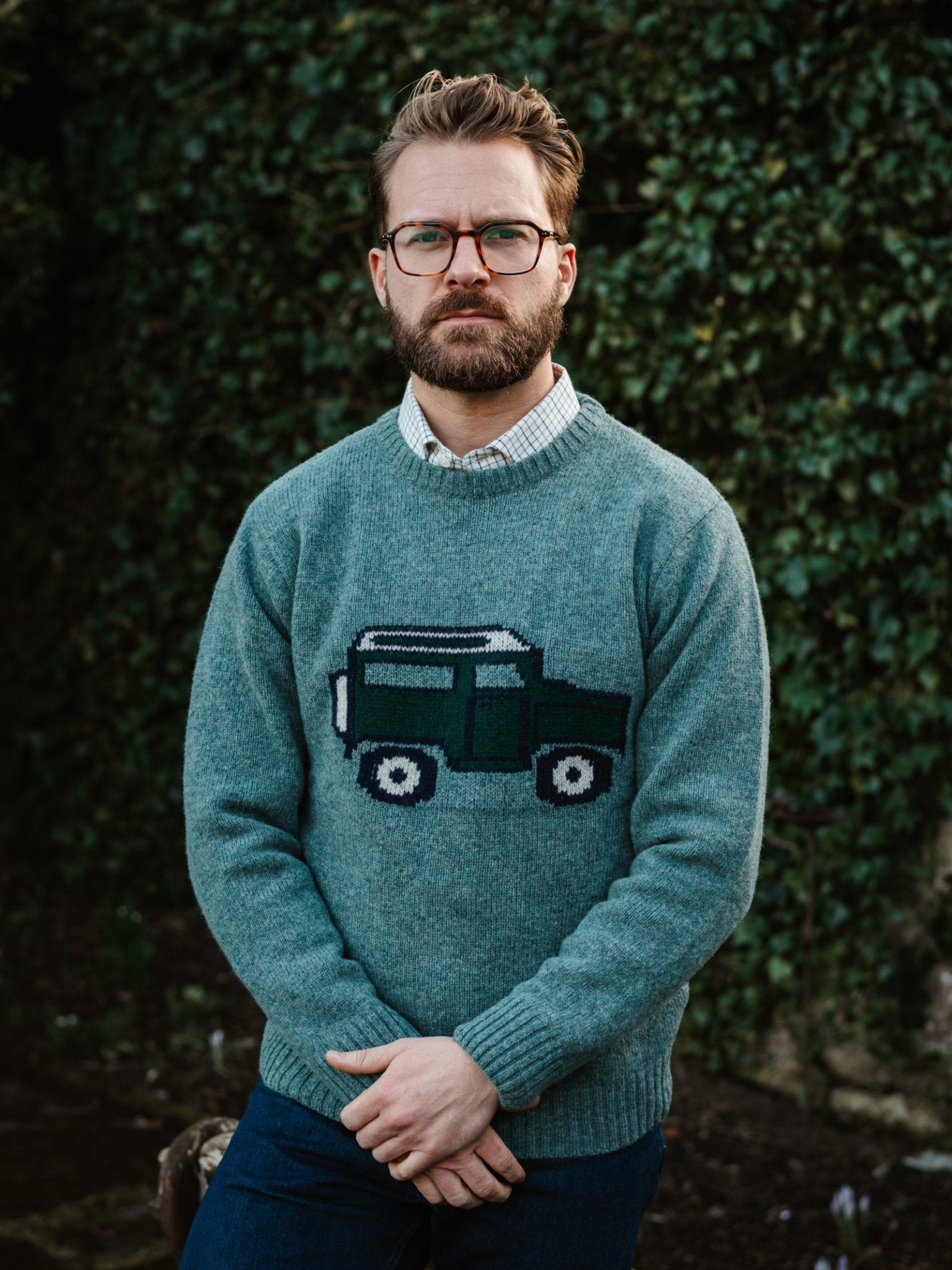 A bearded man with glasses stands outdoors in front of greenery, wearing a Campbell's of Beauly Unisex Land Rover Defender Jumper—a stylish tribute to British motoring—while crossing his hands and looking at the camera.