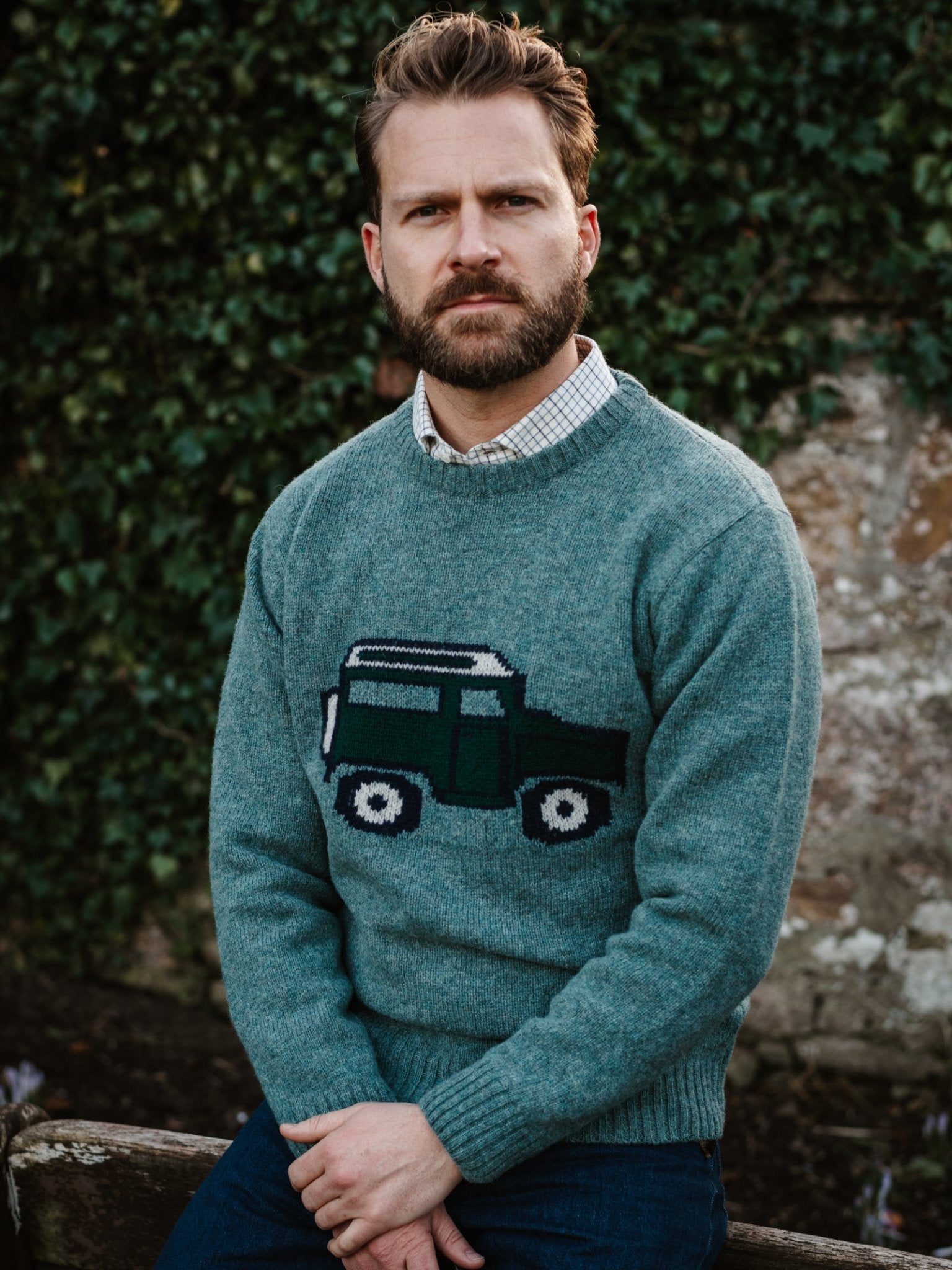 A bearded man with styled hair sits outdoors by ivy-covered stone, wearing the Campbell's of Beauly Unisex Land Rover Defender Jumper over a checked shirt. The green knit features a classic car design inspired by British motoring.