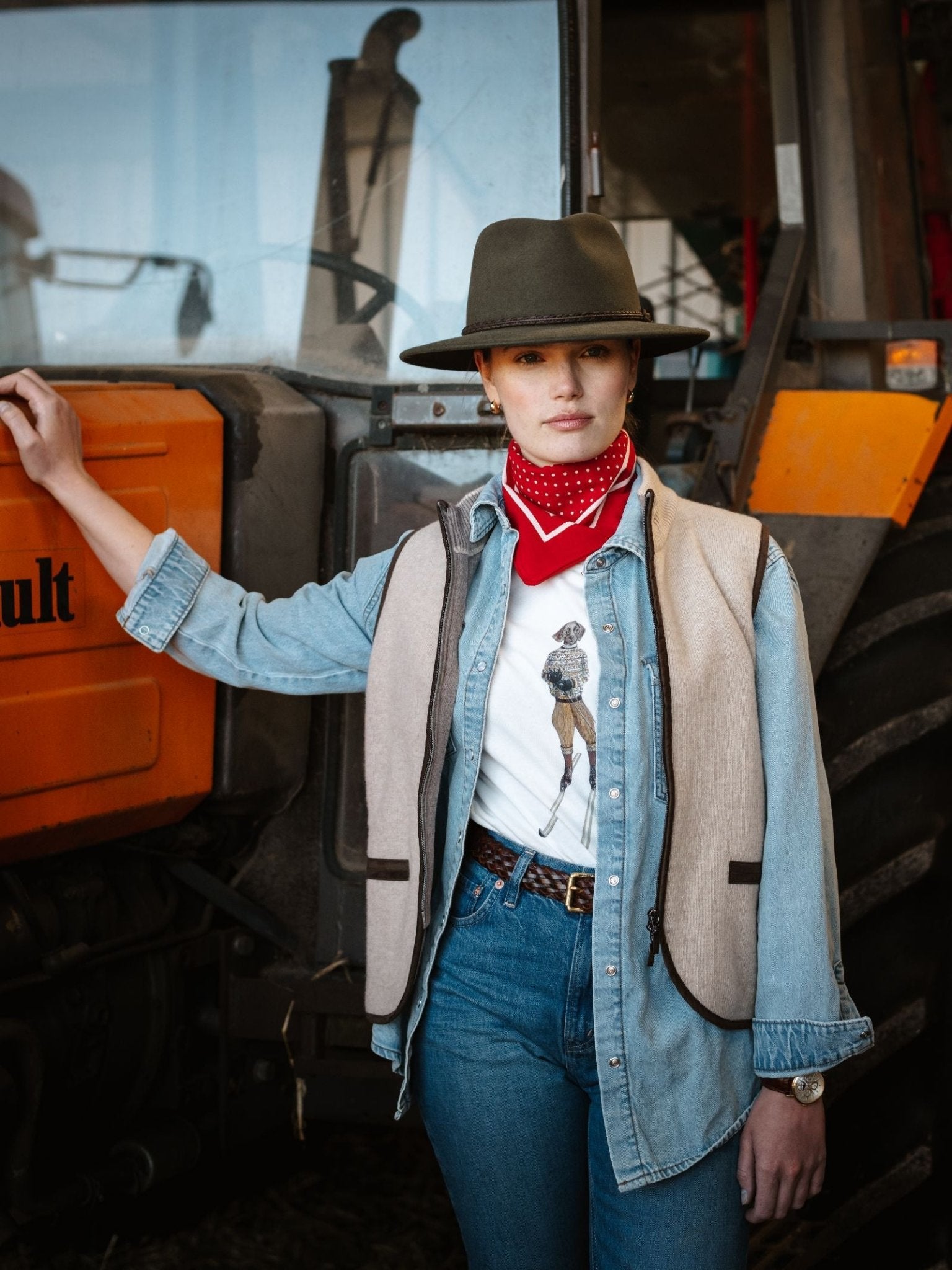 A woman in a denim shirt, jeans, vest, and wide-brimmed hat poses by a large orange tractor. She wears a red bandana and the Campbell's of Beauly Le Ski Labrador T-Shirt, confidently resting one hand on the tractor.
