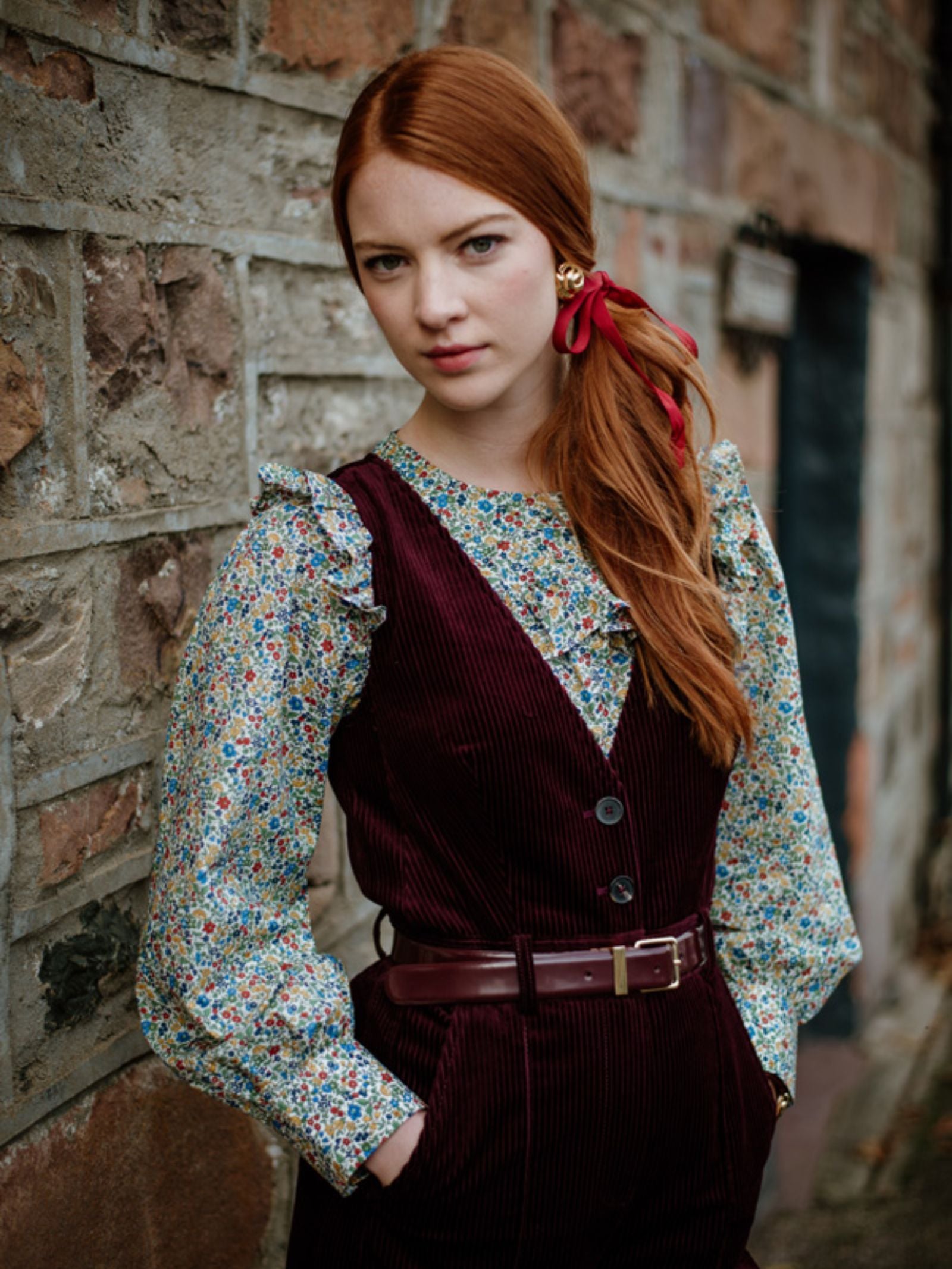 A young woman with long red hair, tied with a red ribbon, stands by a stone wall. She wears Campbell's of Beauly's Auld Stock Liberty Print Peplum Shirt, its floral motif and peplum silhouette beneath a burgundy corduroy vest and matching belt.