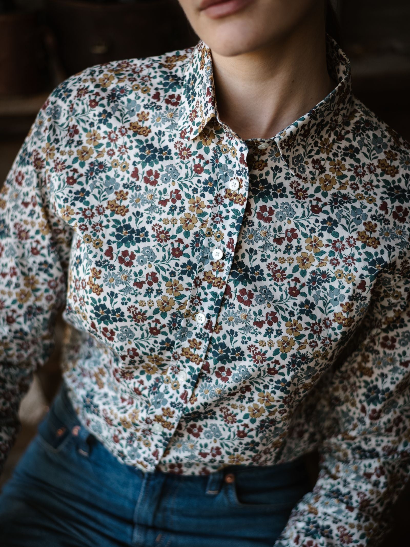 A person wears a collared, long-sleeve Liberty Print Cotton Shirt by Campbell's of Beauly in colorful floral Tana Lawn cotton with dark blue jeans. The image is cropped to show only the lower face and torso.